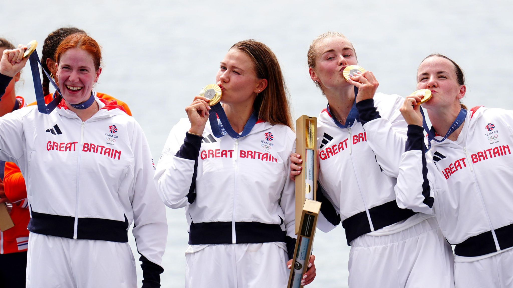 Four women wearing white Team GB tracksuits hold up and kiss their gold circular medals on the waterfront.