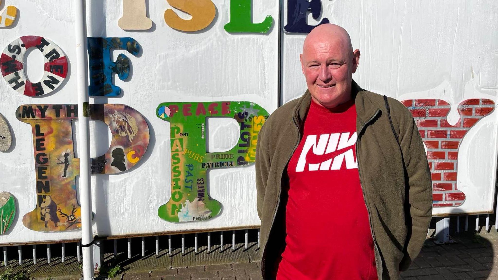 A bald white man in a red Nike t-shirt and brown fleece stands in front of the Isle of Sheppey sign at Sheerness train station