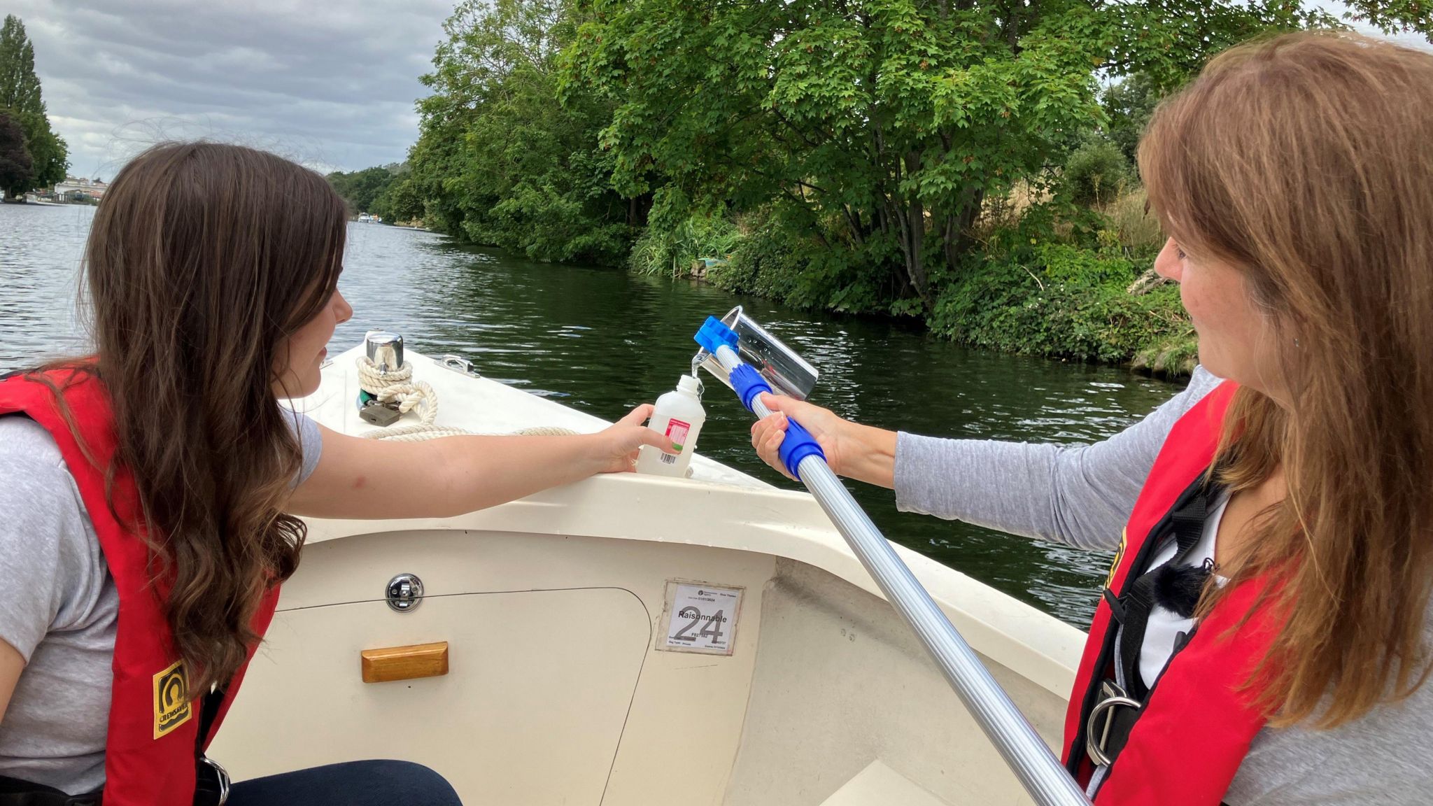 Frances Mateo pouring a river water sample into a container held by Izzy Wittrock while on a boat