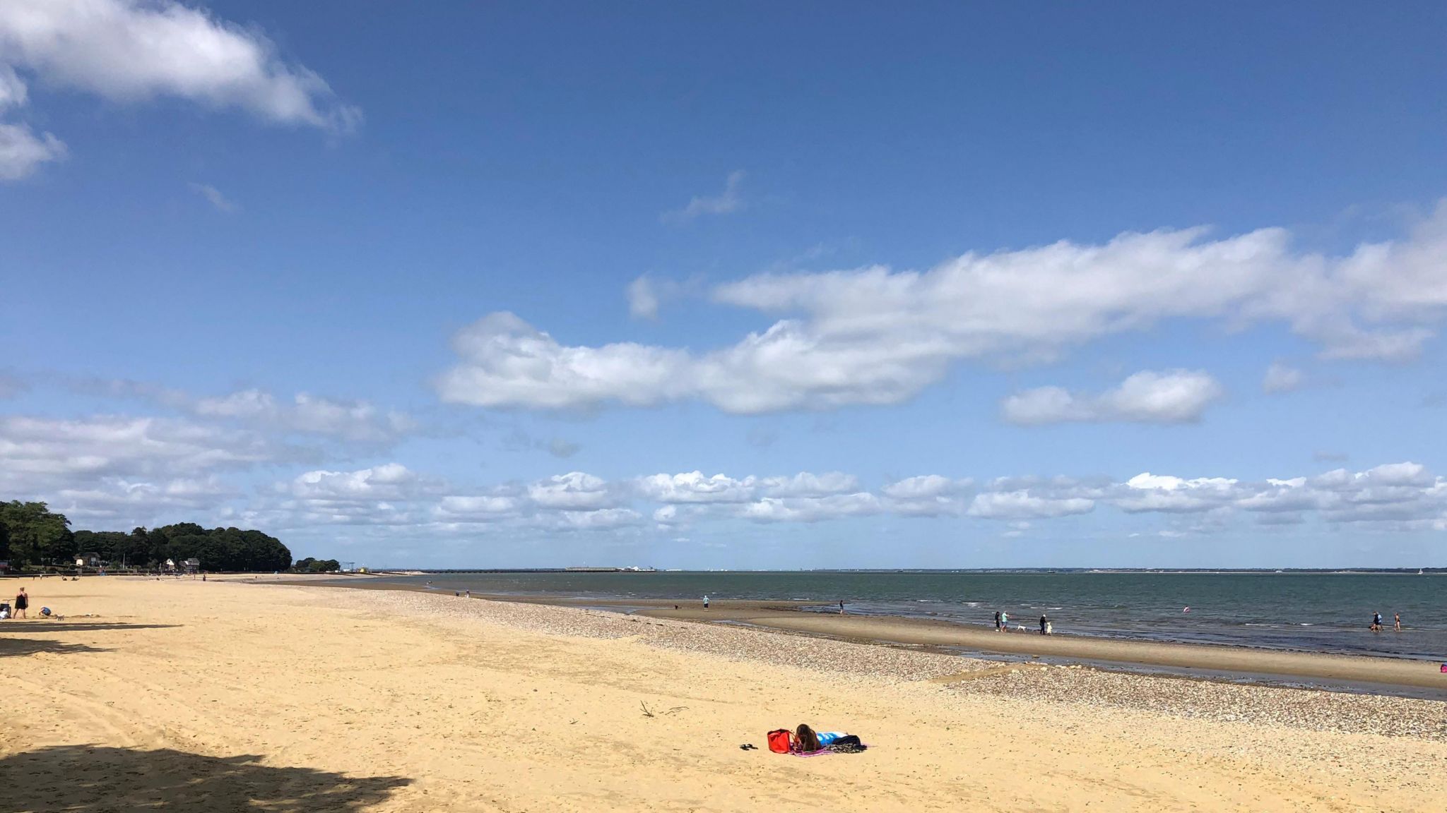 A golden sand beach on a sunny day, there is one person lying down on the beach wearing blue with a red bag. In the distance you can see several people paddling in the sea. Behind the beach you can see green trees and there are several white clouds above in the sky. 