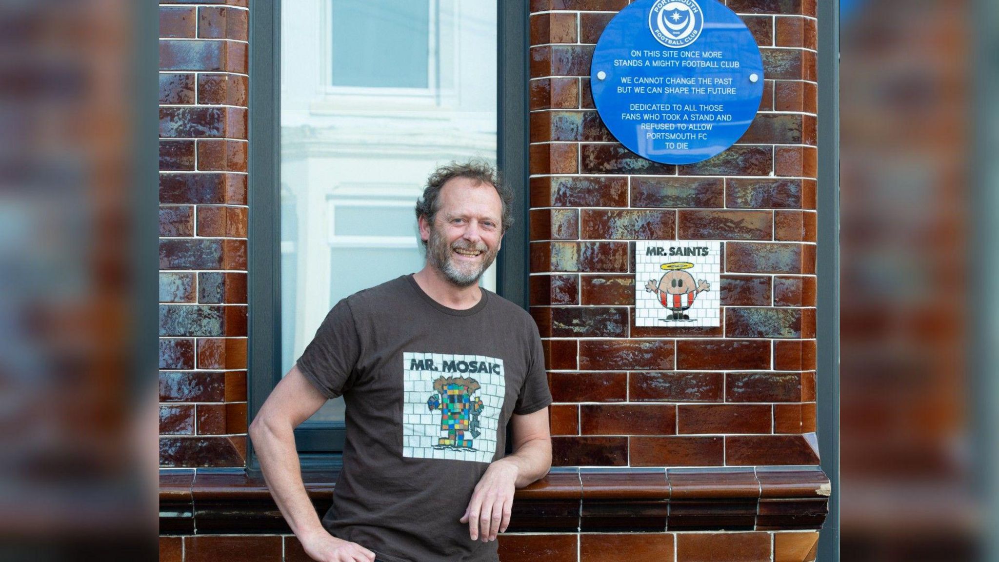 Will Rosie wearing a t-shirt which says "Mr Mosaic" he is leaning on the wall where there is a blue plaque which shows the Portsmouth Football Club crest above text which reads "on this site once more stands a mighty football club. We cannot change the past but we can shape the future. Dedicated to all those fans who took a stand and refused to allow Portsmouth FC to die" underneath the plaque is a white mosaic with a mr man character dressed in a southampton fc kit, it says "mr saints" at the top