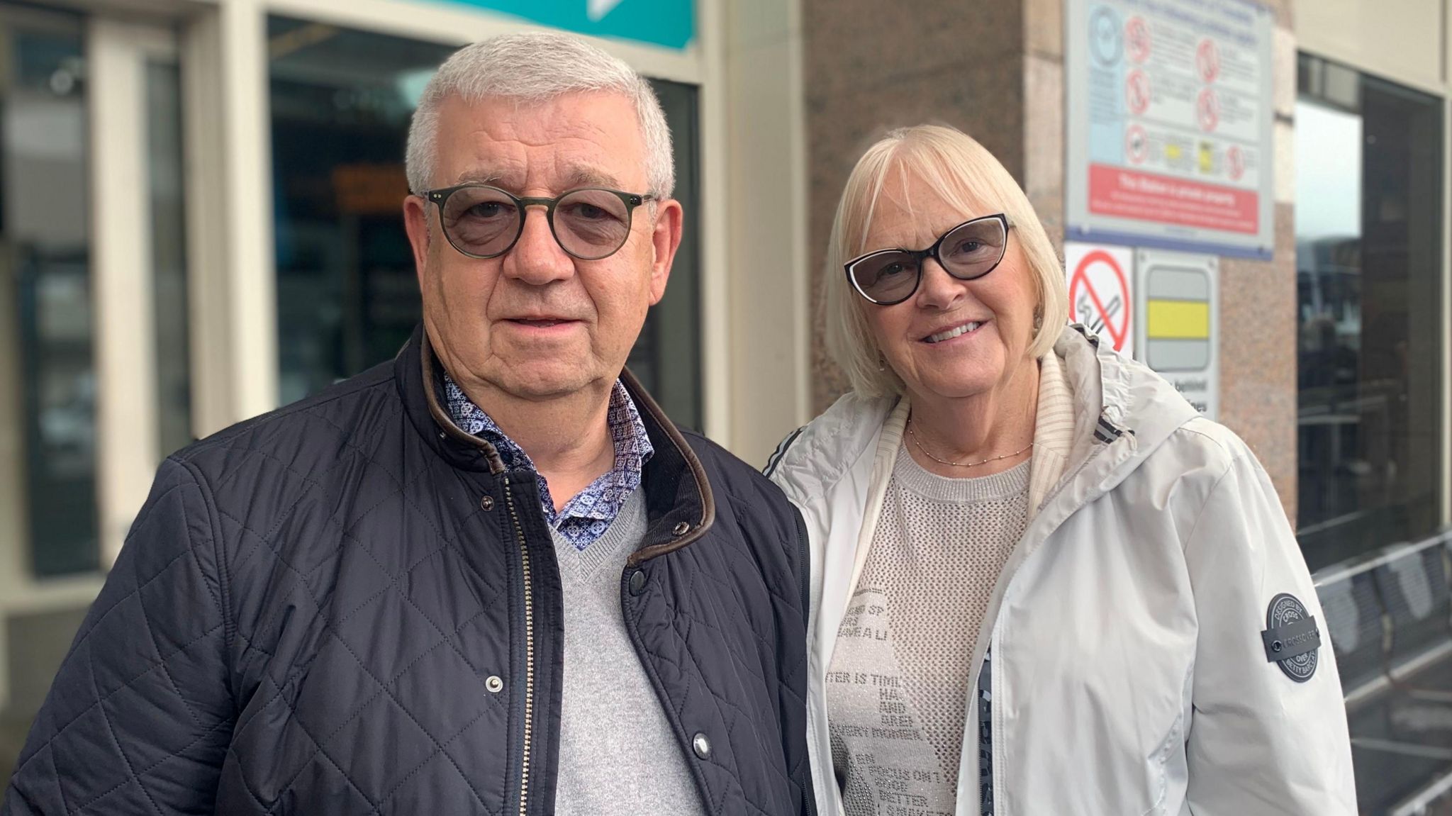 Derek and Anne Smyth - a grey-haired man wearing circular glasses and a navy quilted jacket stands beside a blonde woman wearing glasses and a grey raincoat