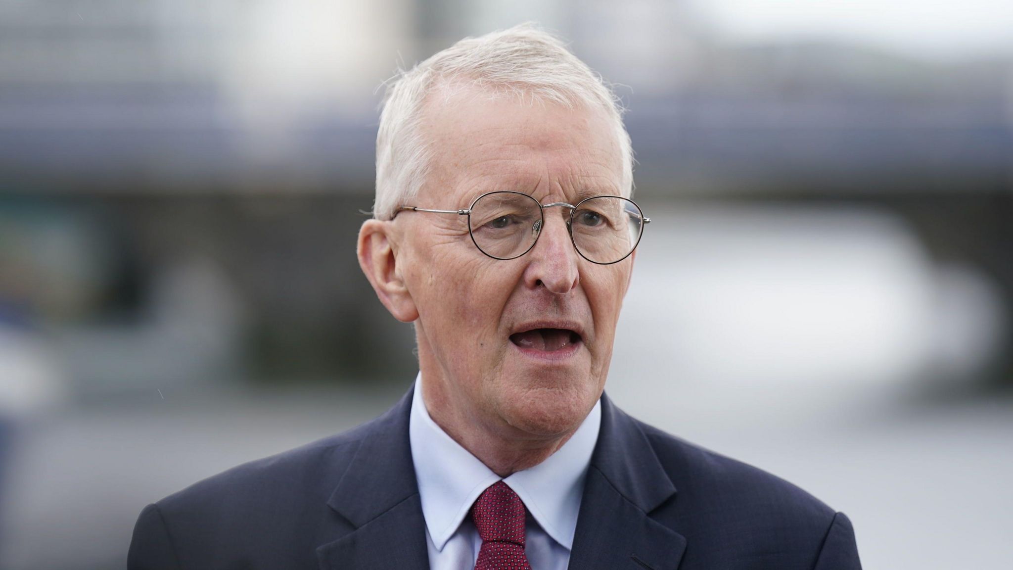 A man with white hair and black circular glasses wears a dark suit jacket, a light shirt and red tie