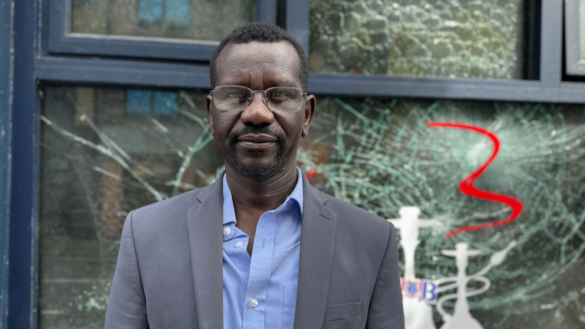 Man with black hair, black beard and glasses wearing a grey suit jacket and light blue top standing in front of a smashed window 