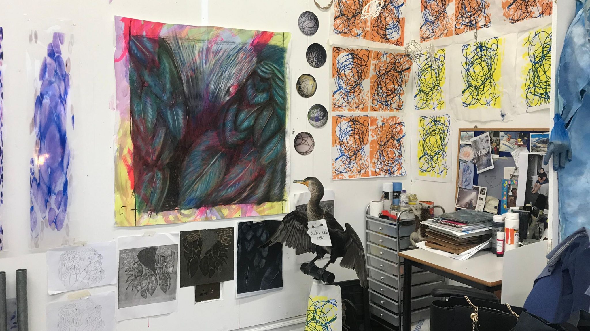 A picture of the inside of Joanne's studio. The room contains a small desk and a set of drawers, but the walls are covered in abstract, brightly coloured pictures and there is what appears to be a large taxidermy bird in the centre on a stand.  