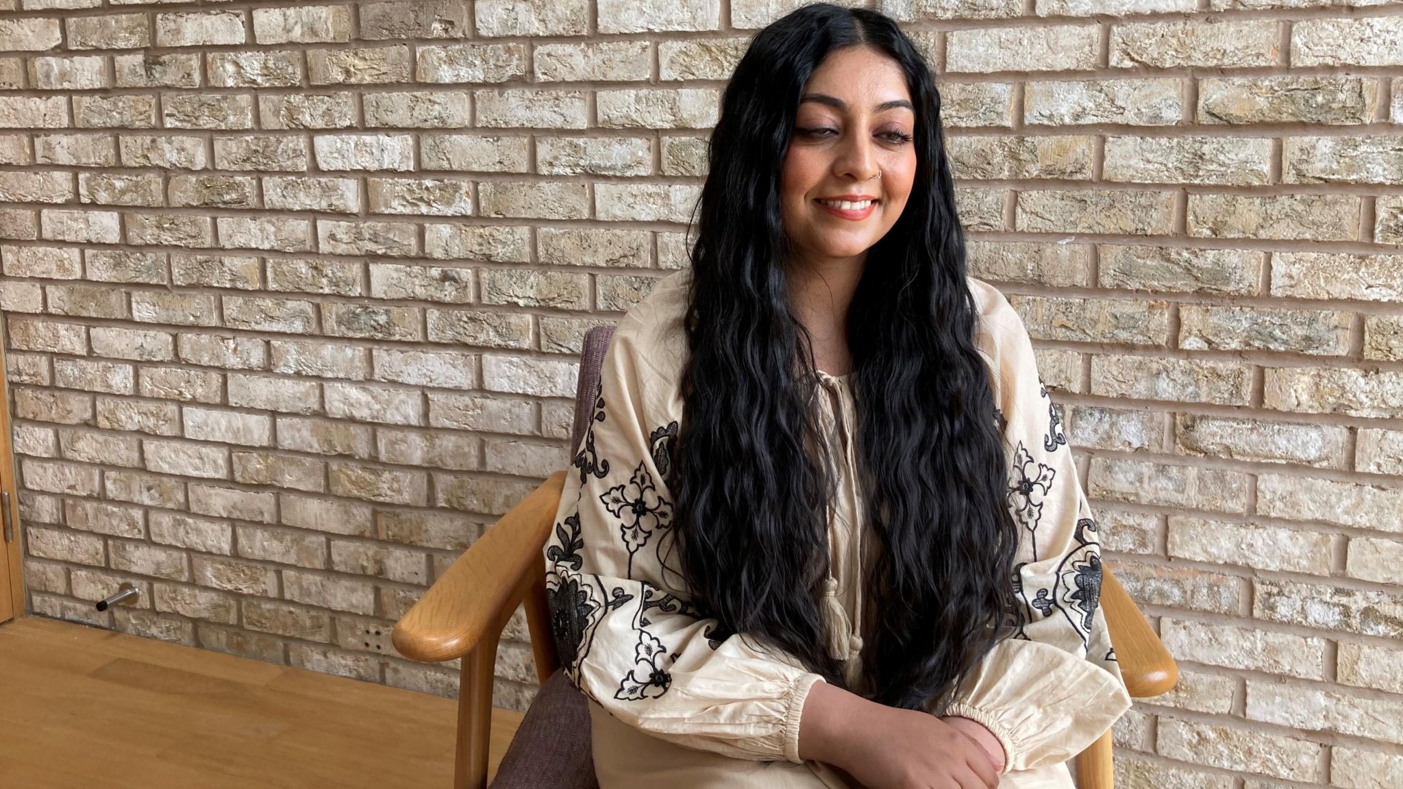 Waseema Shaikh wears a white blouse with blue embroidered floral detailing, sitting in a wooden chair with a brick wall in the background 