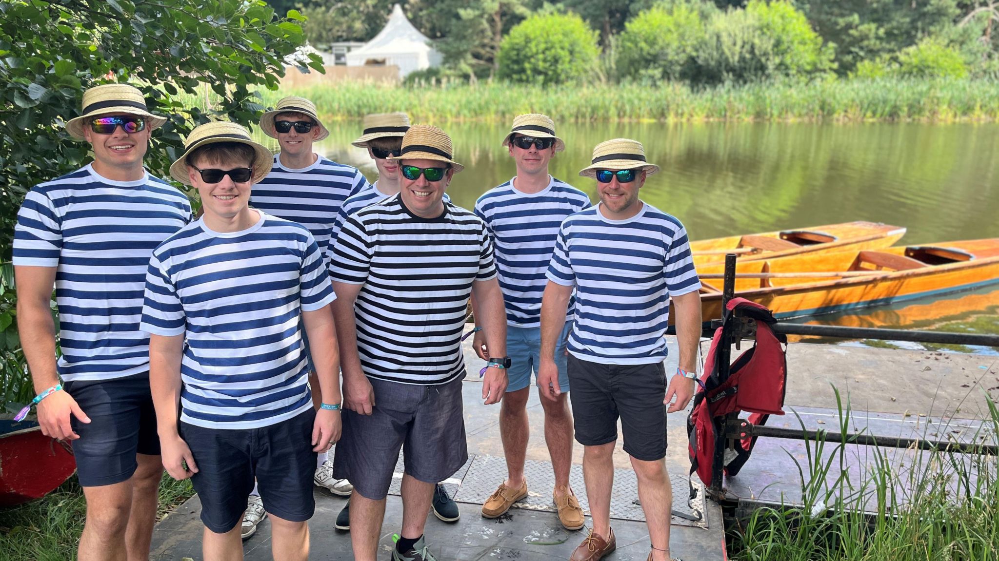 Seven men in straw boater hats and blue and white striped tops stand by punts on the lake at Henham park, Southwold