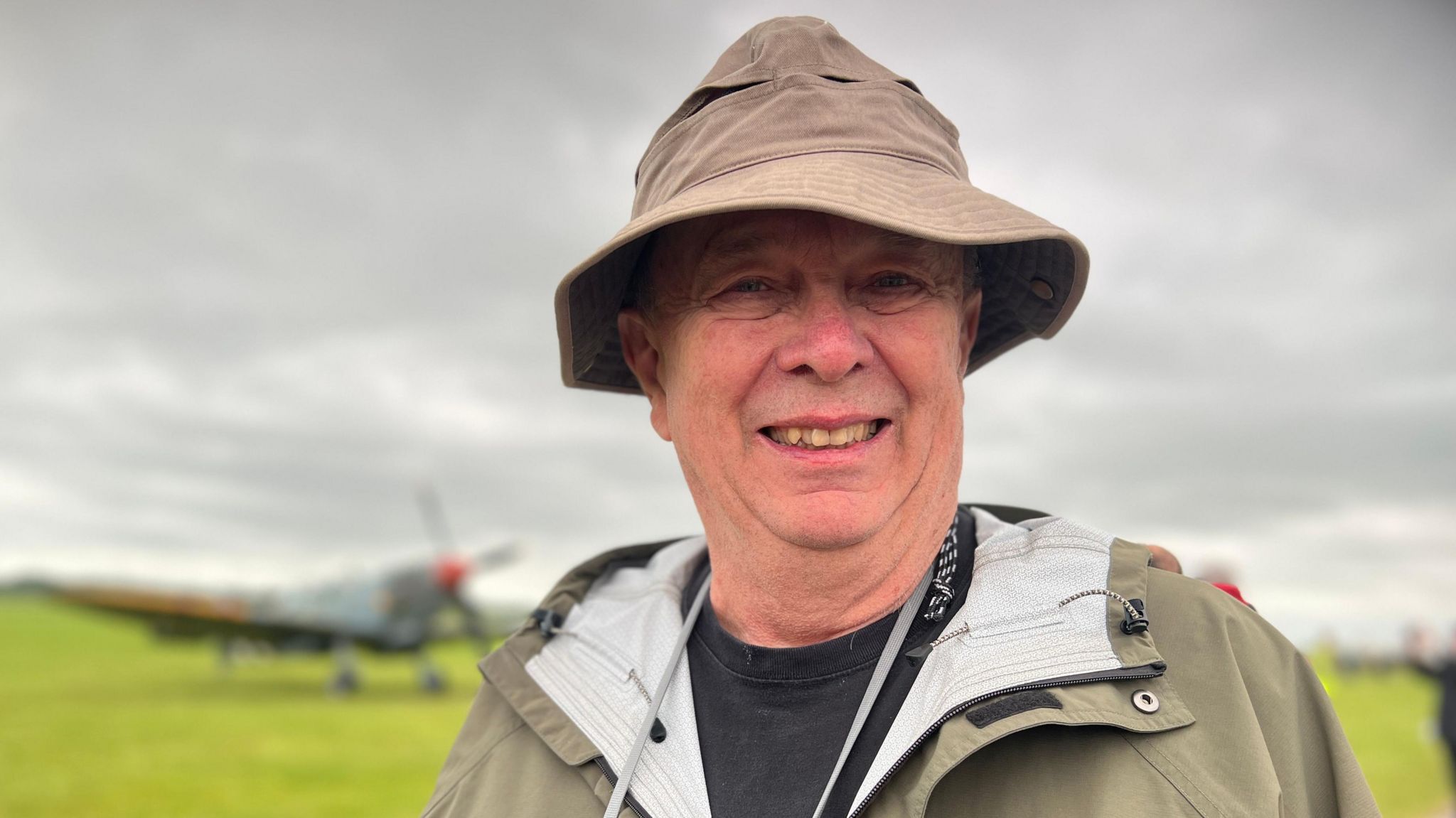 Donald Johnson wearing a beige hat and coat with the airfield and an aircraft behind him, Duxford