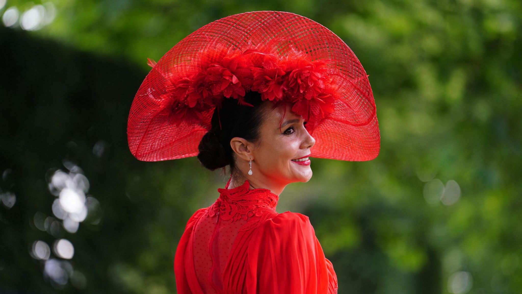 Woman looks away from the camera wearing a large red feathered headdress, with her dark hair in a low slick bun and a high collared red dress