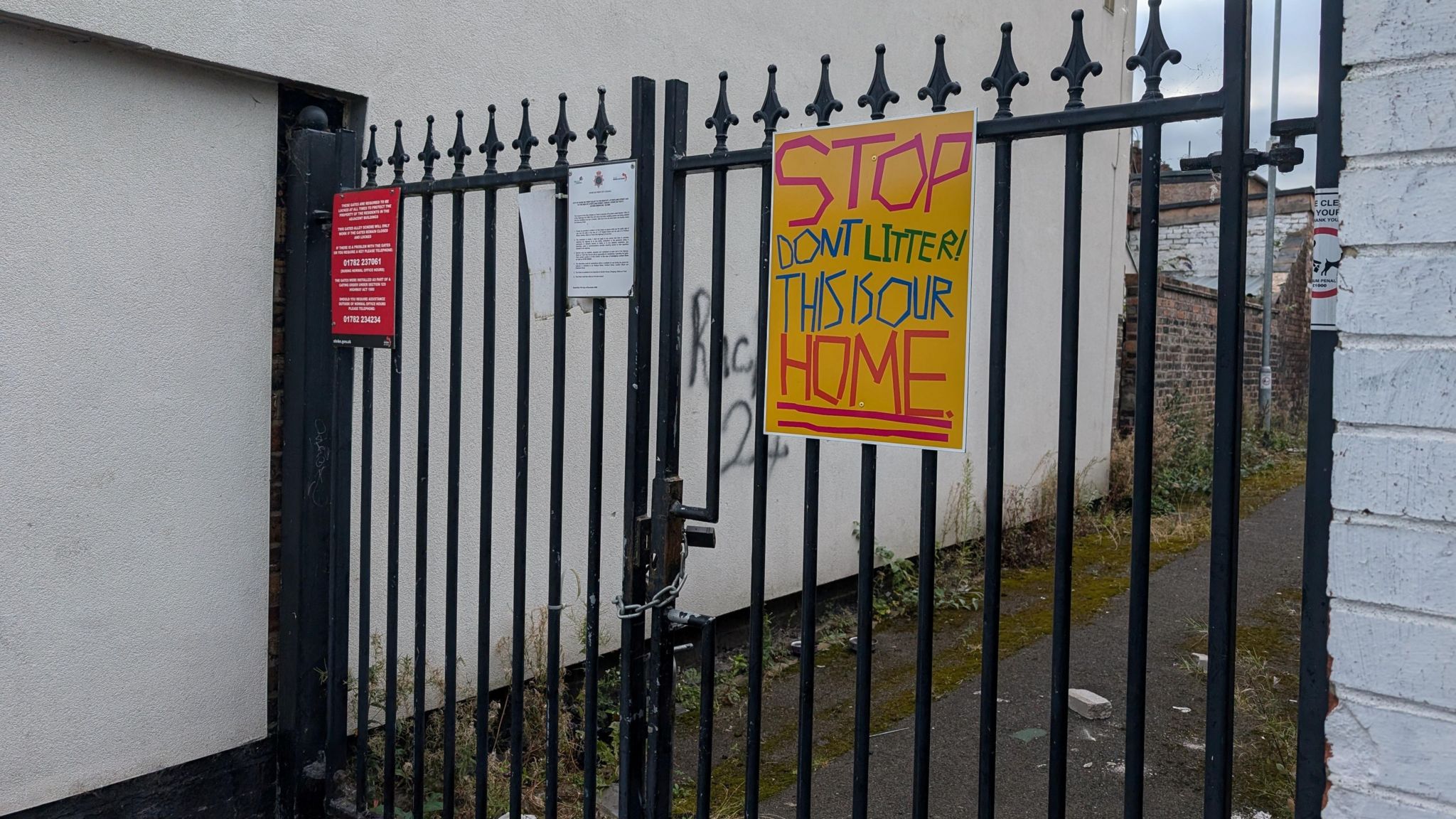 A black gate in an alleyway with a yellow sign attached to it that says "stop, don't litter, this is our home".