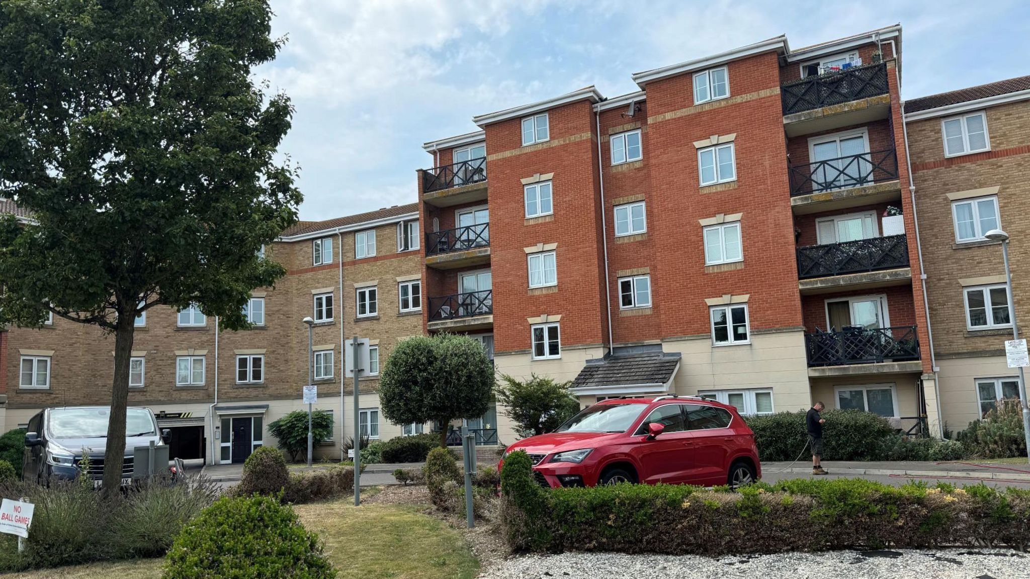 A block of flats, with a red car parked outside beside a small garden