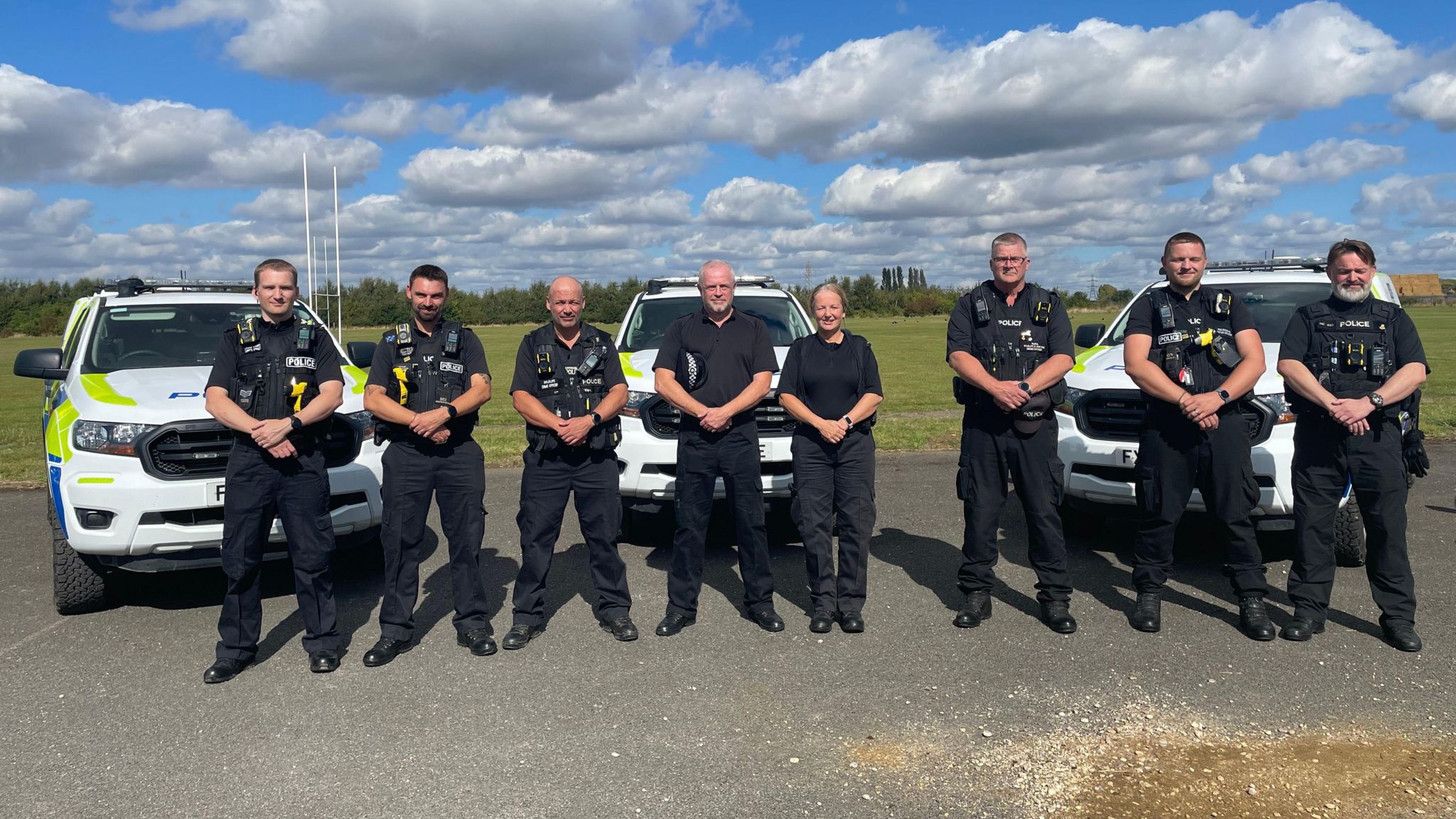 Eight police officers, all wearing black uniforms, stand in front of three all-terrain police cars, parked at the edge of a sports field.