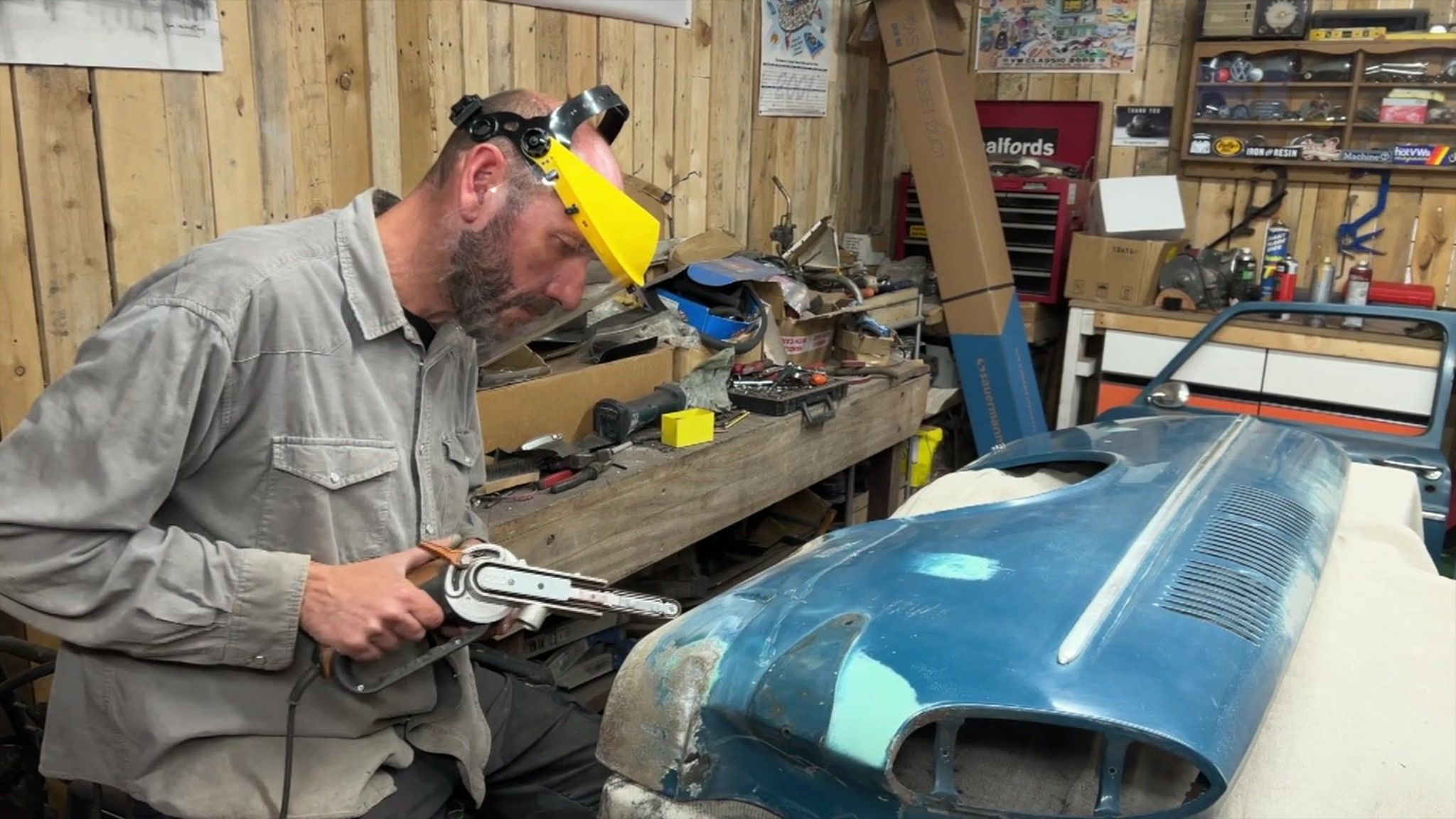 Rob Gwinnett wearing a yellow safety visa works on part of a VW vehicle inside a workshop. It has wood panelled walls and shelves filled with mechanical parts. He is wearing a grey shirt and has a dark beard and dark hair with a bald spot on top of his head.