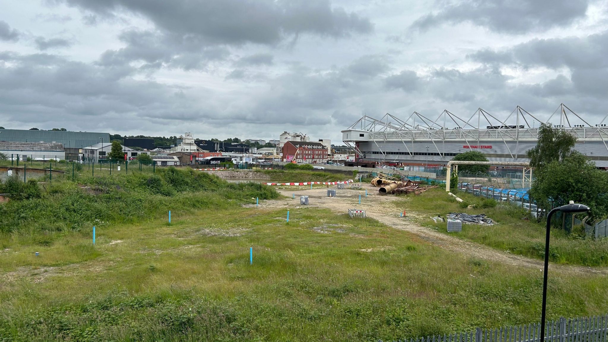 An empty plot of land with a few bits of debris littered across it and some sparse vegetation, with St Mary's stadium in the background