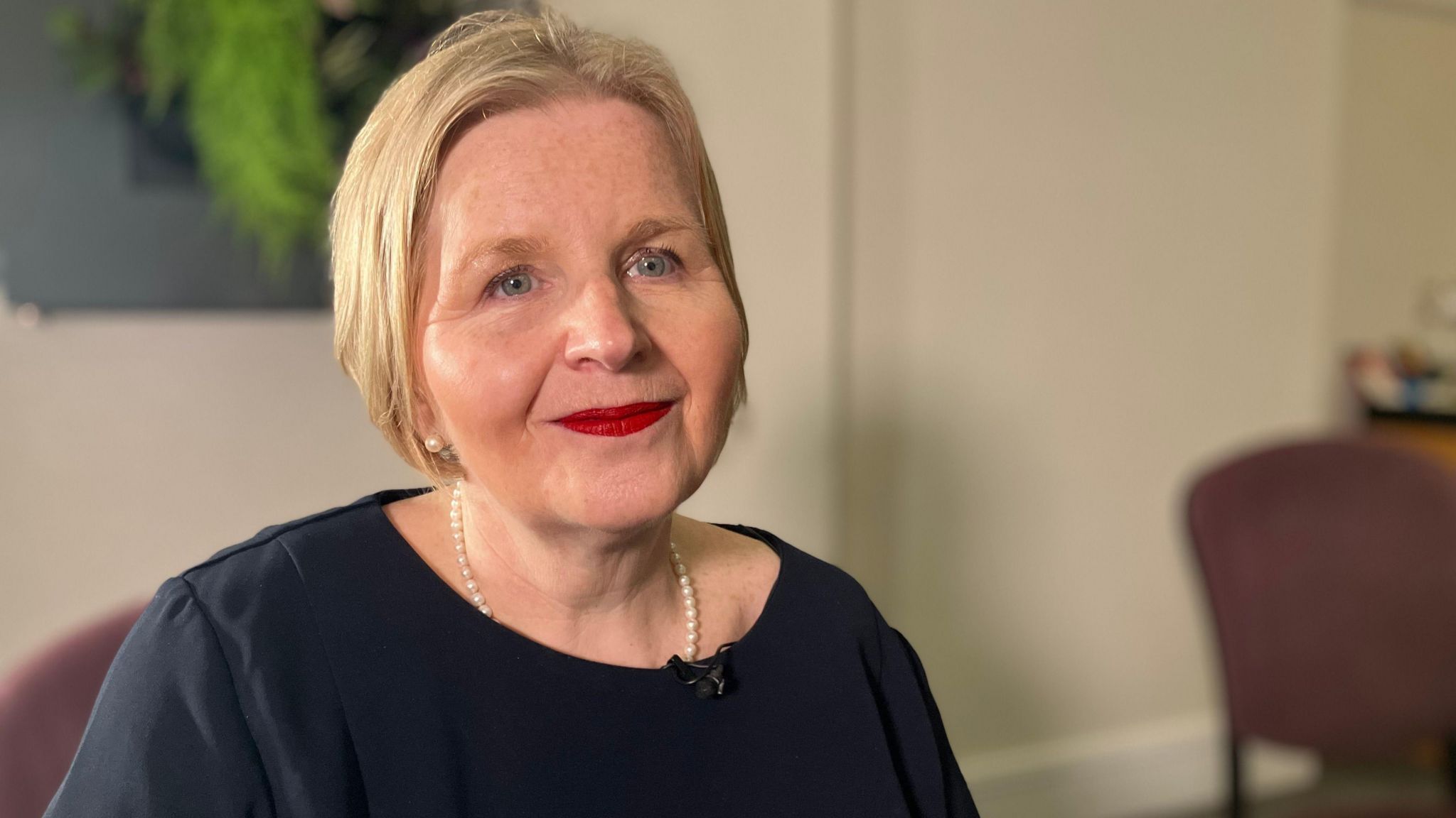 Donna Ockenden sits in a chair as she looks off to the right. She has blonde hair in a short bob and is wearing a dark top, pearl necklace and earrings, and a small clip-on microphone.