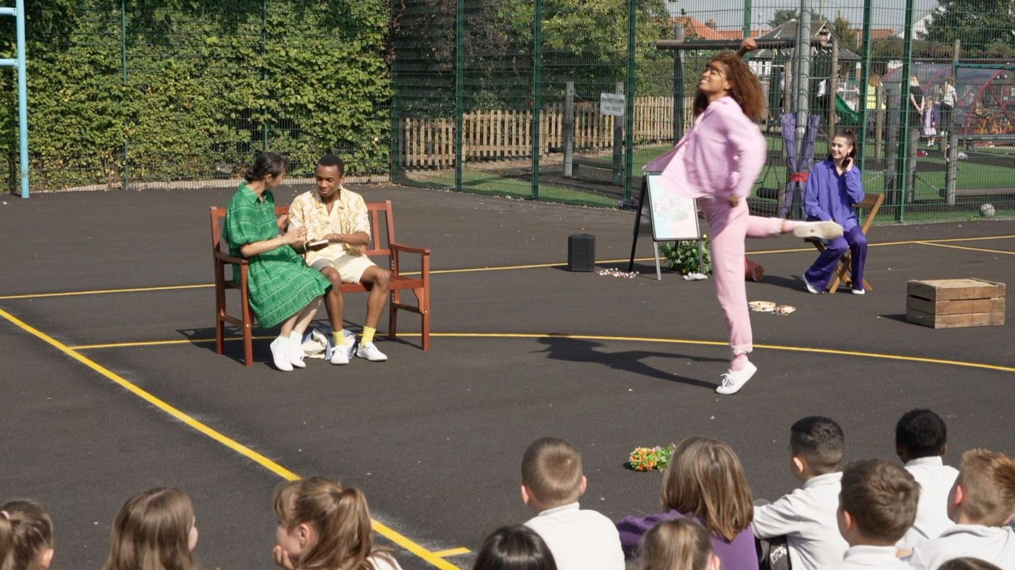 The backs of heads of primary school children as they sit outside on the school playground watching a dance performance. In the background, a man and woman sit on a bench while another woman in pink tracksuit and white trainers does ballet