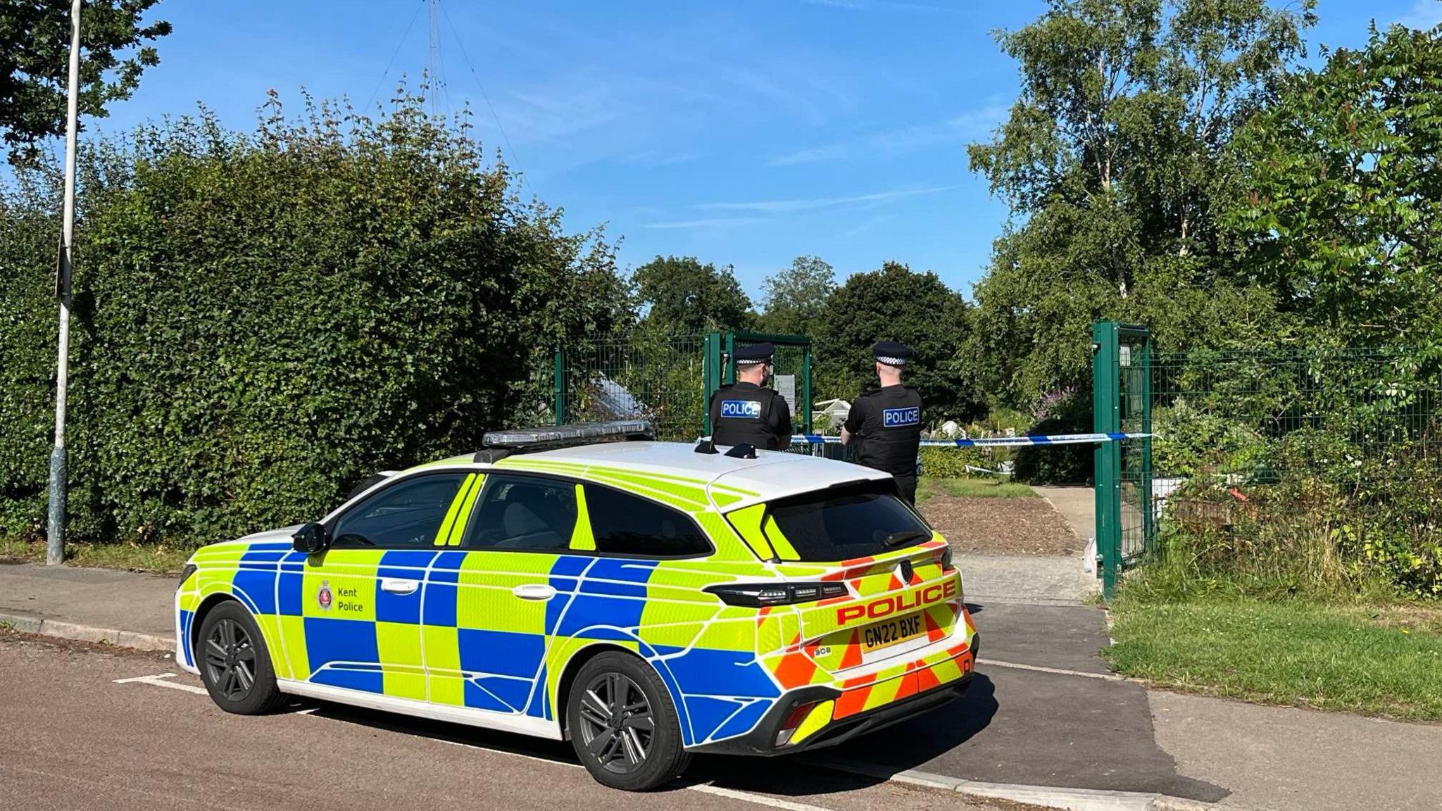 A police car outside the entrance to the allotments with two police officers standing near it