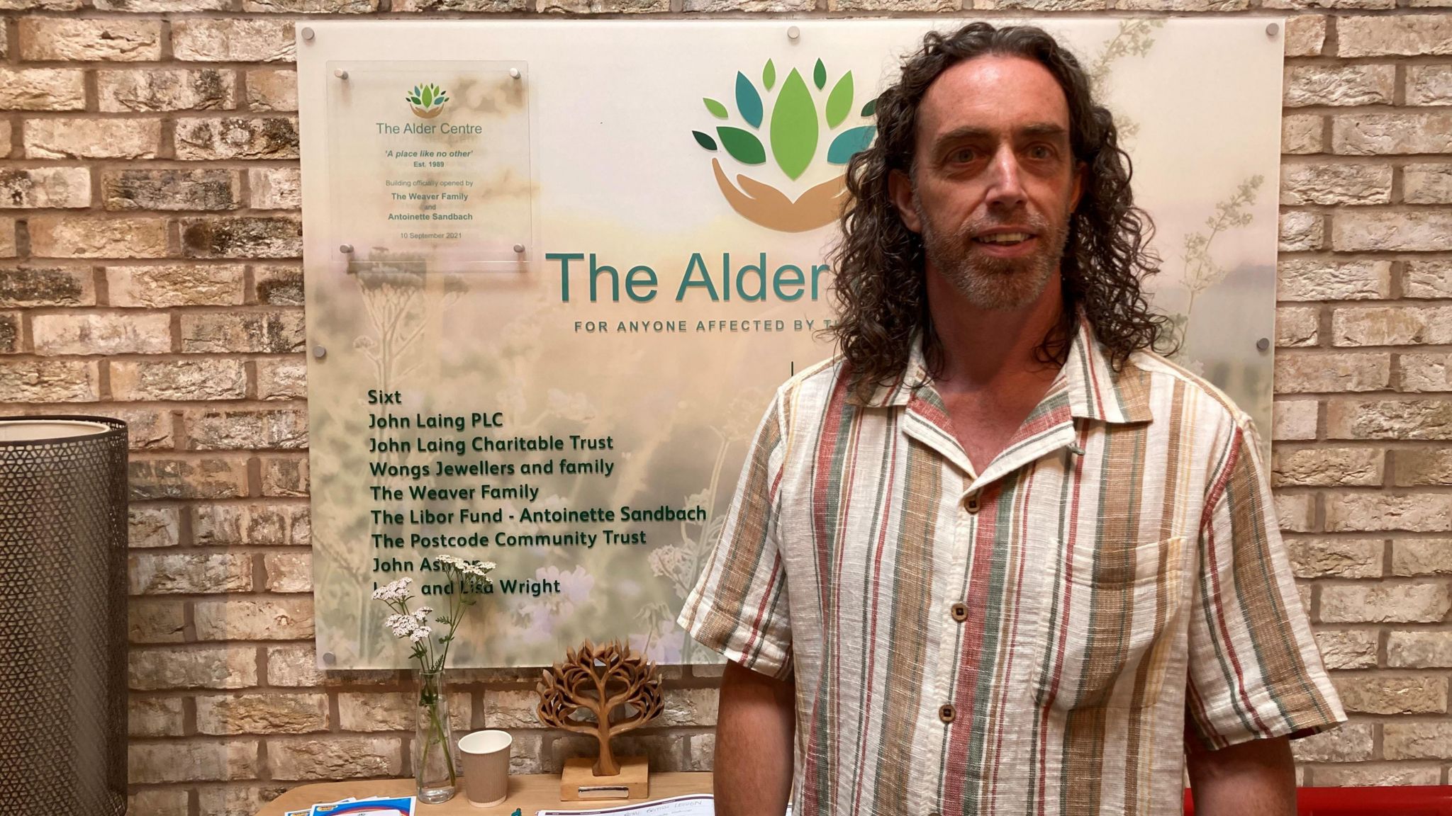 Simon Riley wears a striped cream, red, brown and green short sleeved linen shirt, smiling in front of a sign for the the Alder Centre