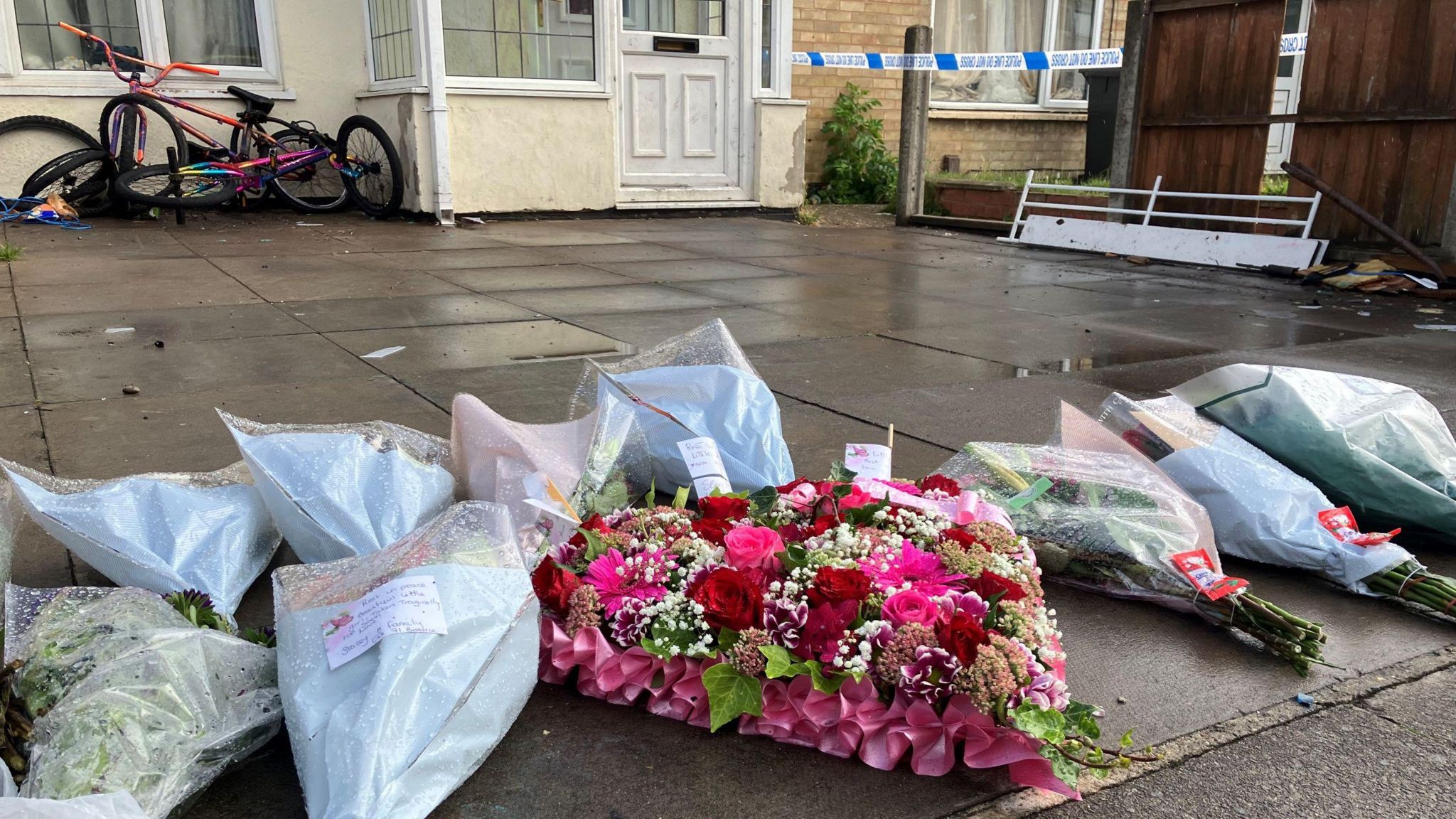 Ten wreaths of flowers left outside a house, in the background children's bikes and police tape can be seen 