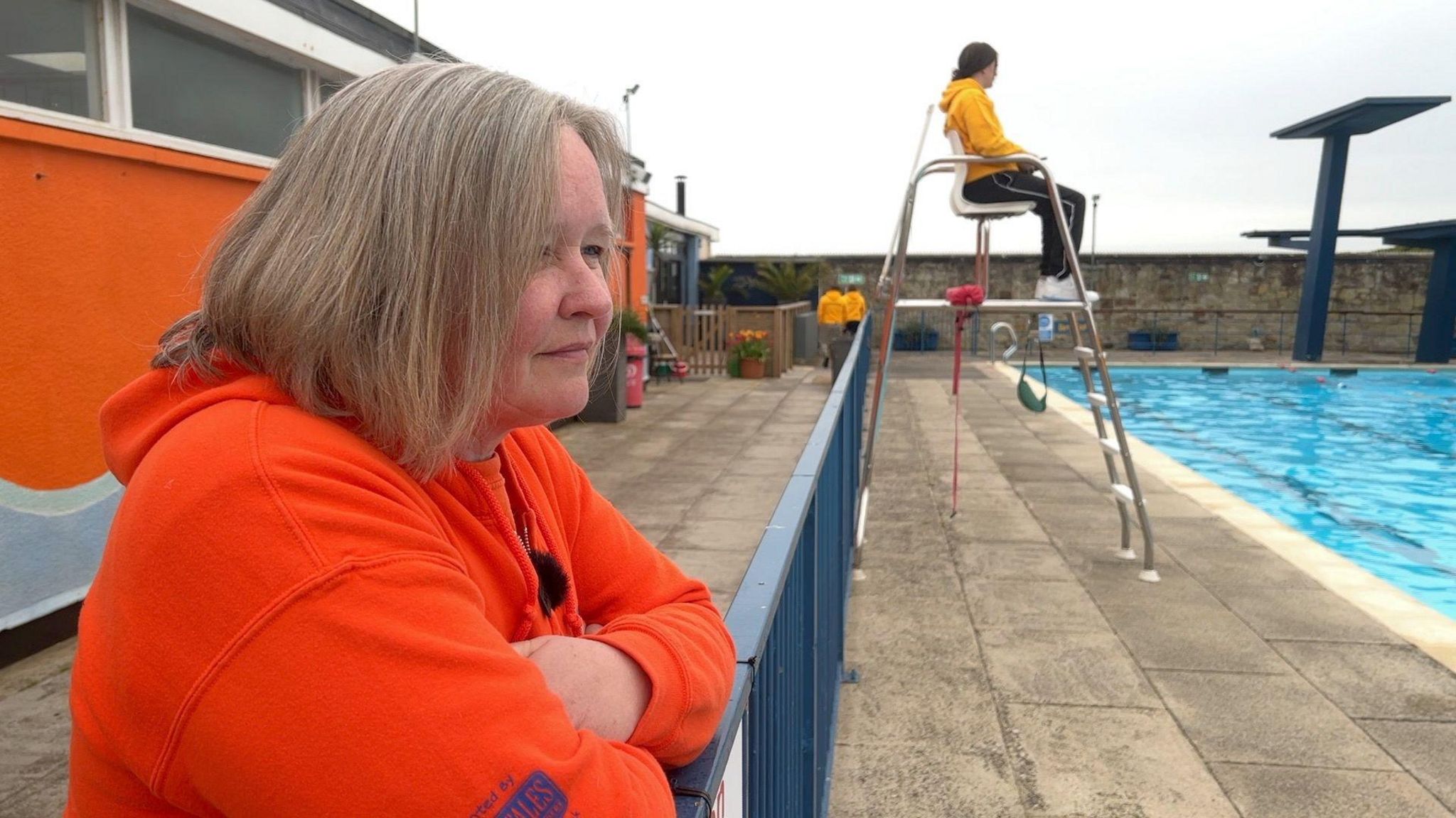 Emma Pusill looks at a swimming pool to her right. She is leaning on a blue railing