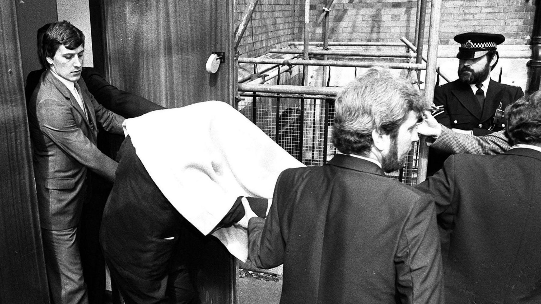 Black and white image showing two police detectives wearing suits escorting a man who is bent over and concealed beneath a blanket from a  court building. A bearded police officer in uniform looks on.