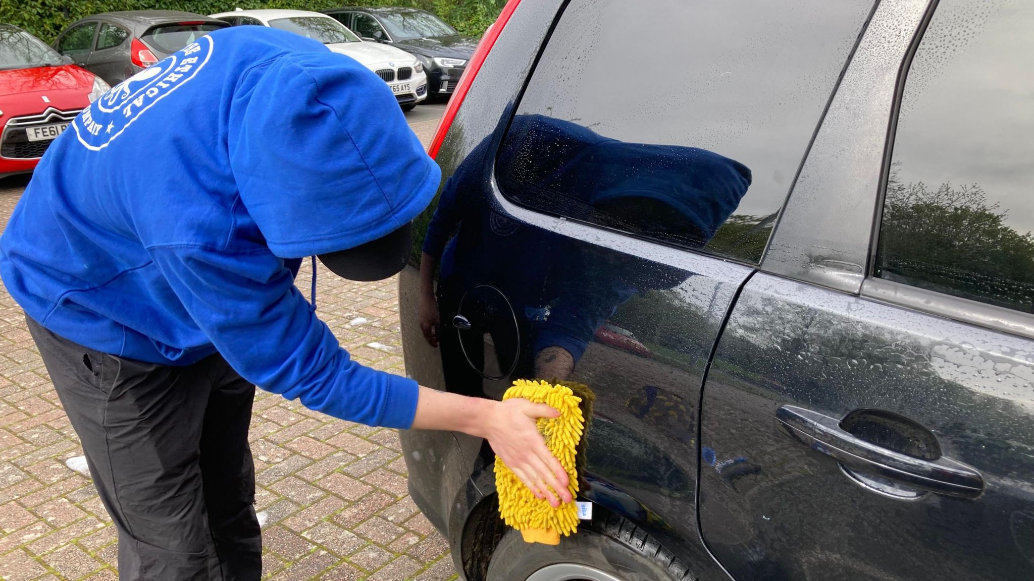 A man in a blue hoodie washing the back of a dirty car