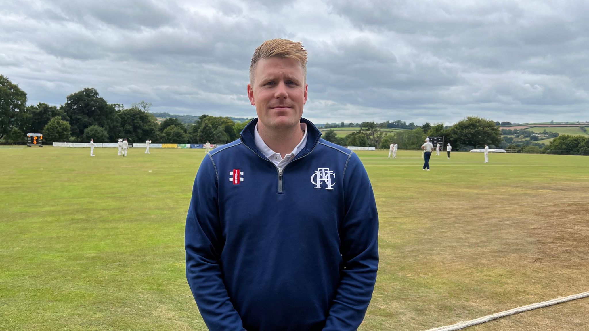 Pete Randerson facing the camera with his hands clasped in front of him. Behind him cricketers play a game on the green. 