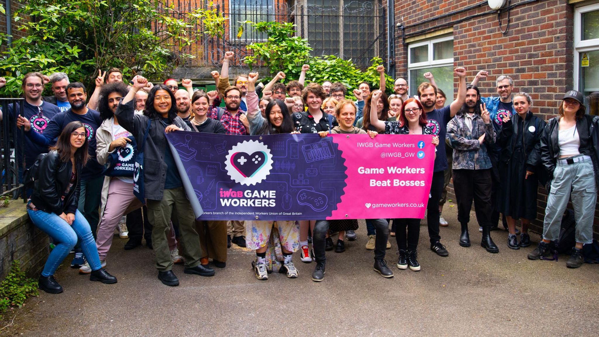 A group of about 30 casually dressed people standing in the courtyard of an office building. Four people at the front are holding a purple and pink banner with the IWGB union Game Workers' branch logo and the slogan "Game Workers Beat Bosses". Some members of the group have their clenched fists raised above their heads.