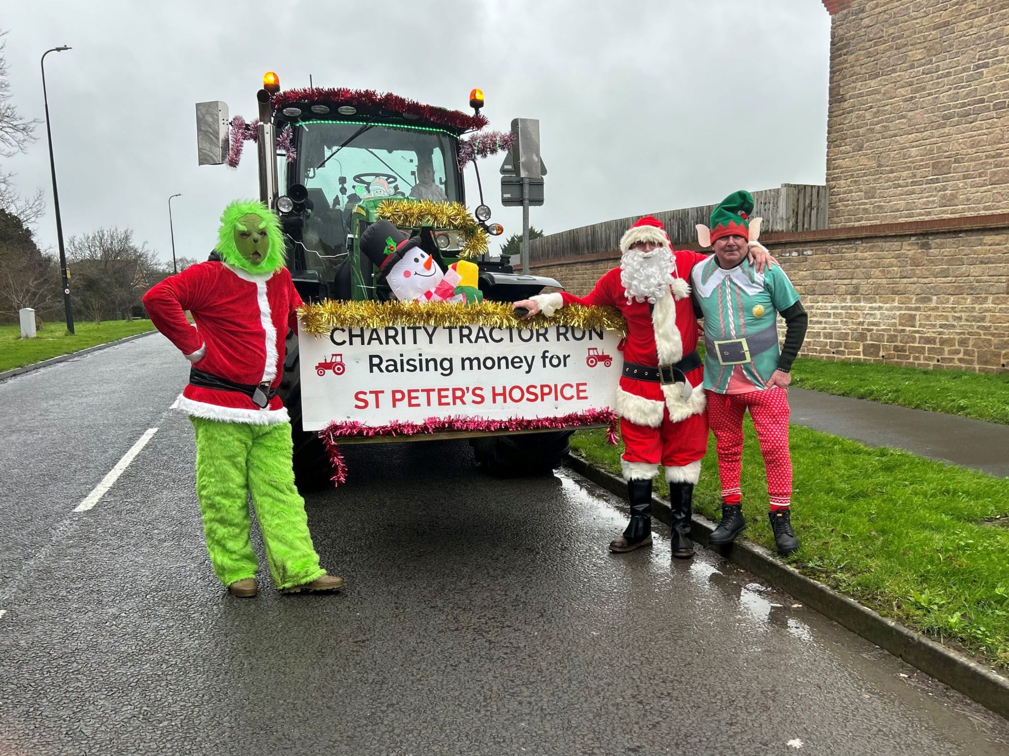 Three men dressed as the Grinch, Father Christmas and an elf are standing near a tractor which says Charity Tractor Run Raising money for St Peter's Hospice