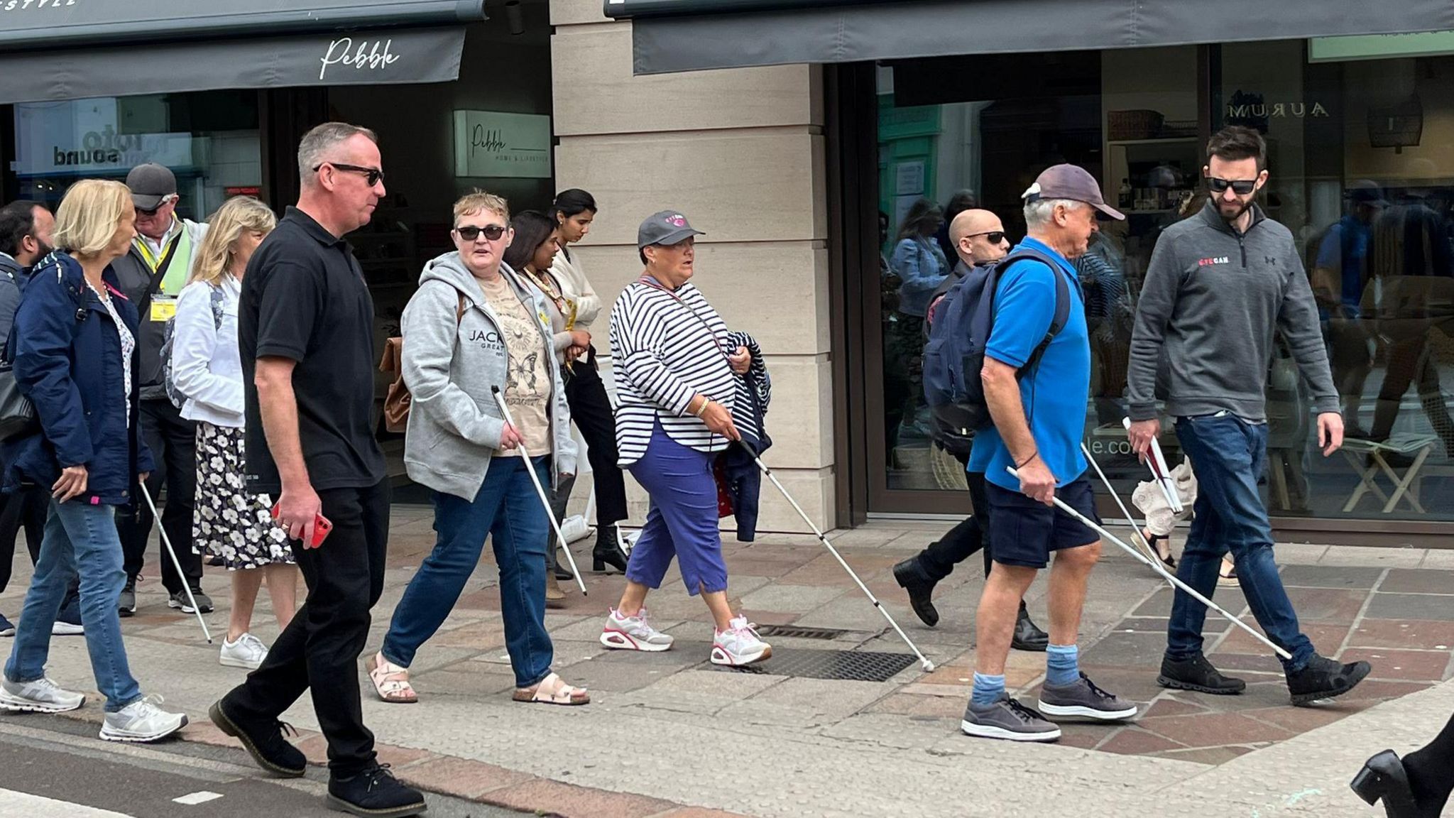 A mixed group of about 12 adult men and women walking on a wide pavement outside a shop, some of whom are using white canes.