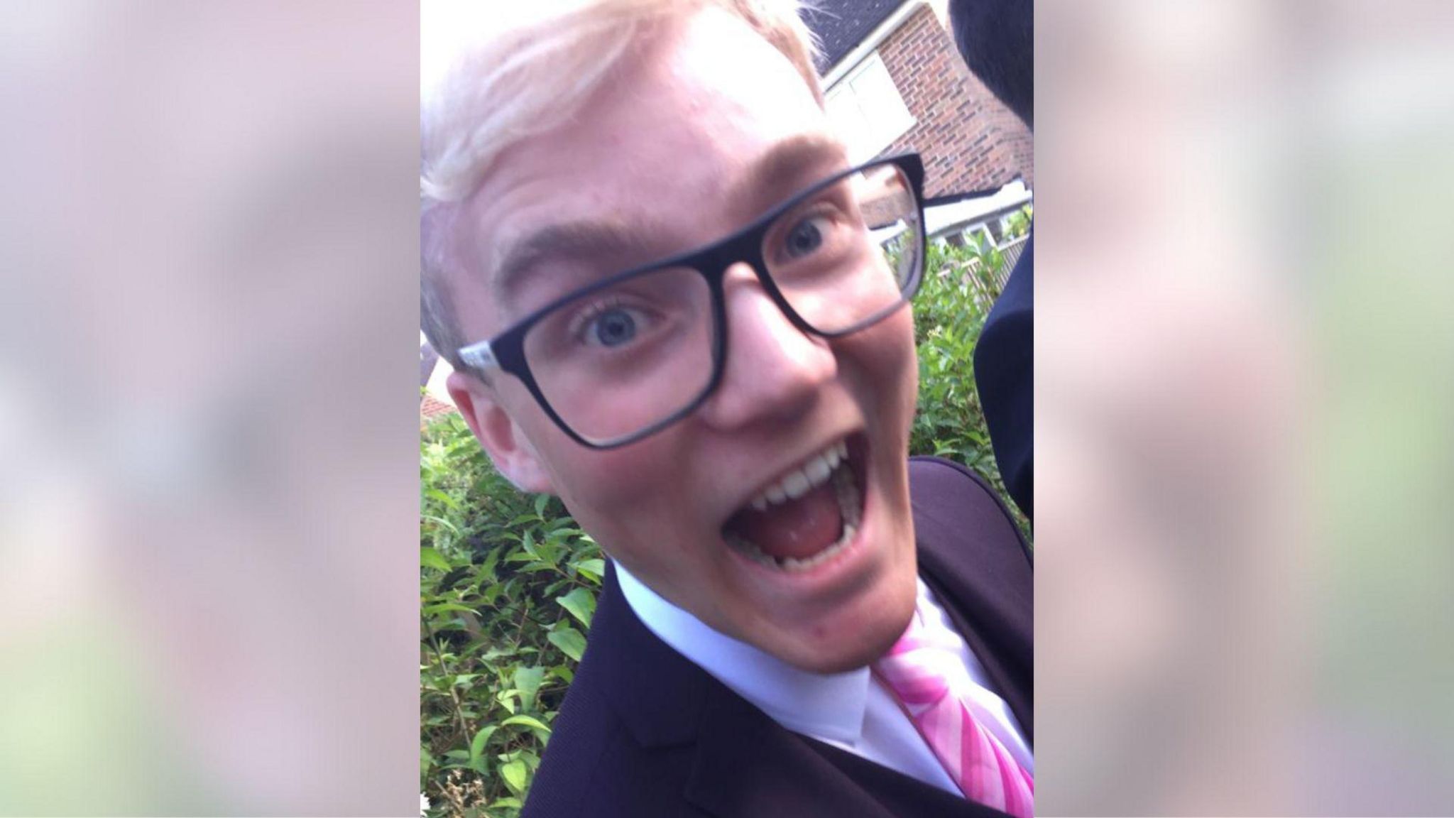 A man with blonde hair and glasses. He is stood in front of a bush wearing a white shirt, pink tie and black blazer. He is smiling with an open mouth.