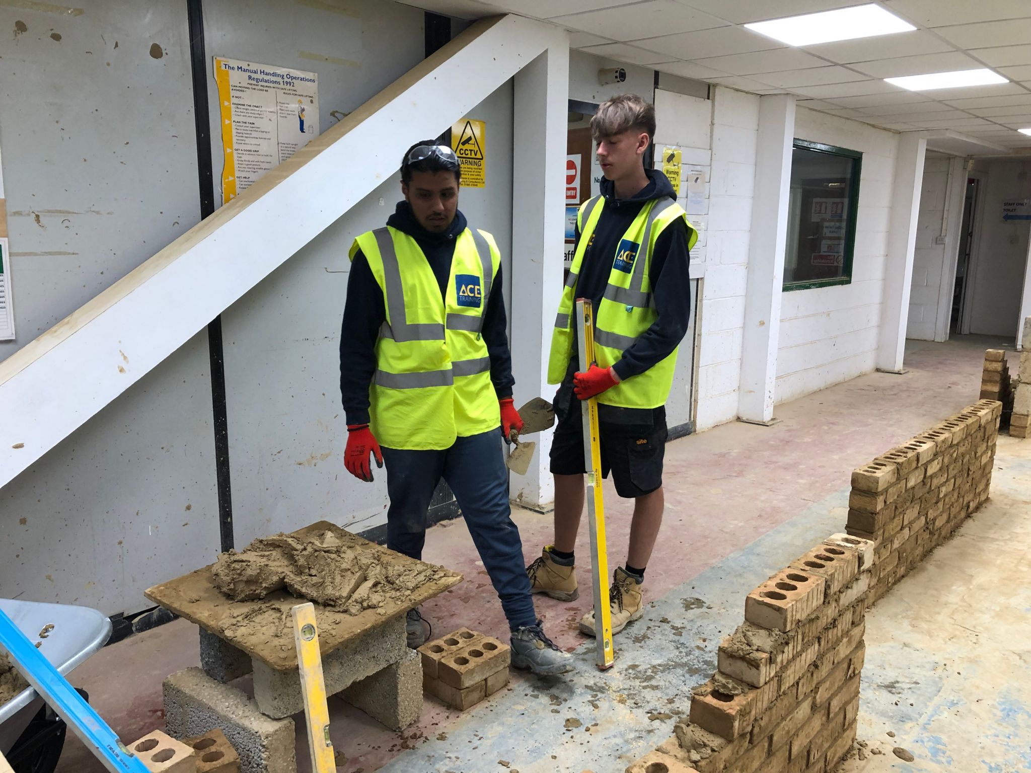 Two of the boys stand in a workshop wearing high-vis jackets and practice their bricklaying skills