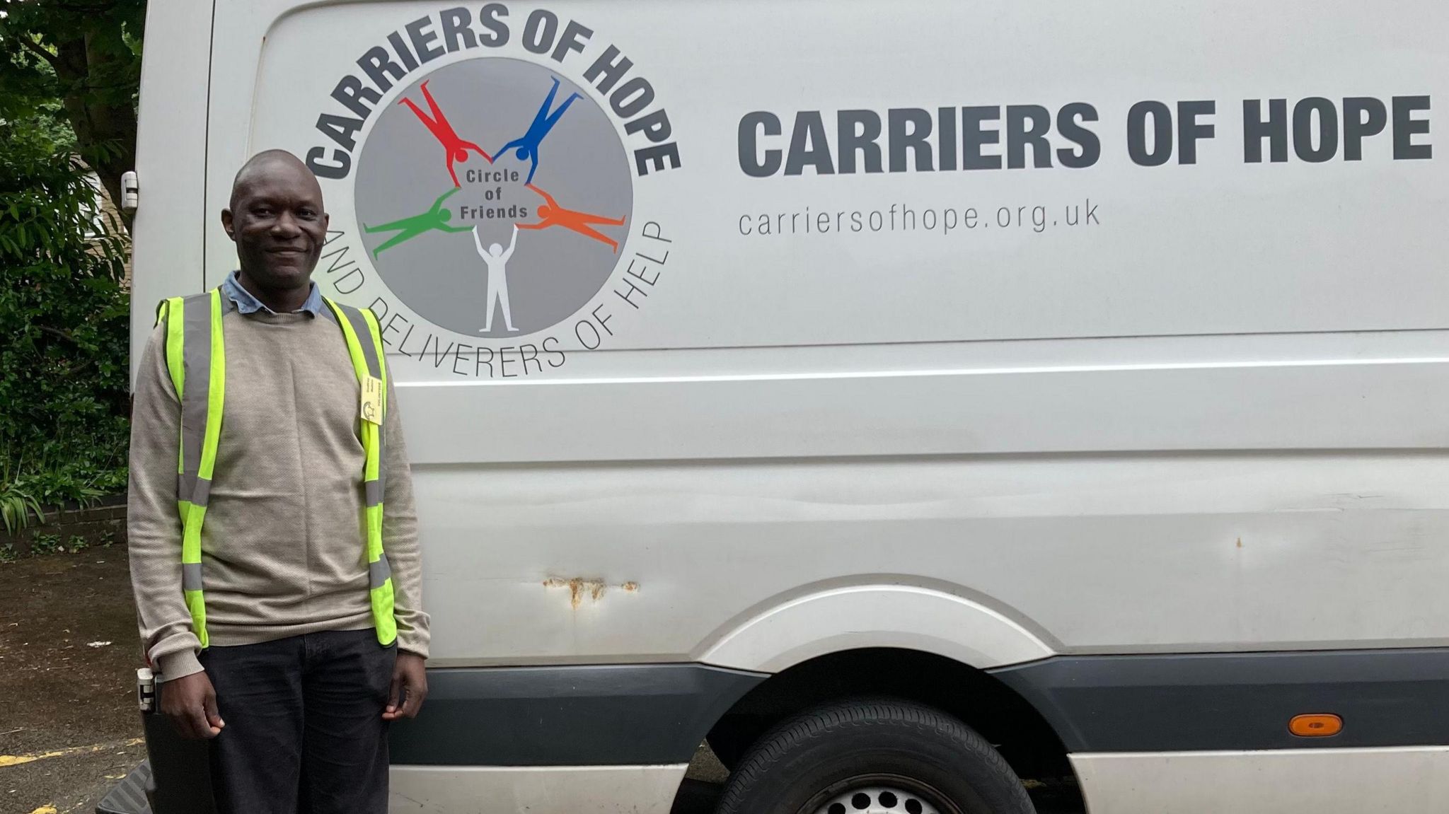 Godfrey, a black man in a beige top and a hi-vis jacket, stands before a white van with the words "Carriers of Hope" written on it