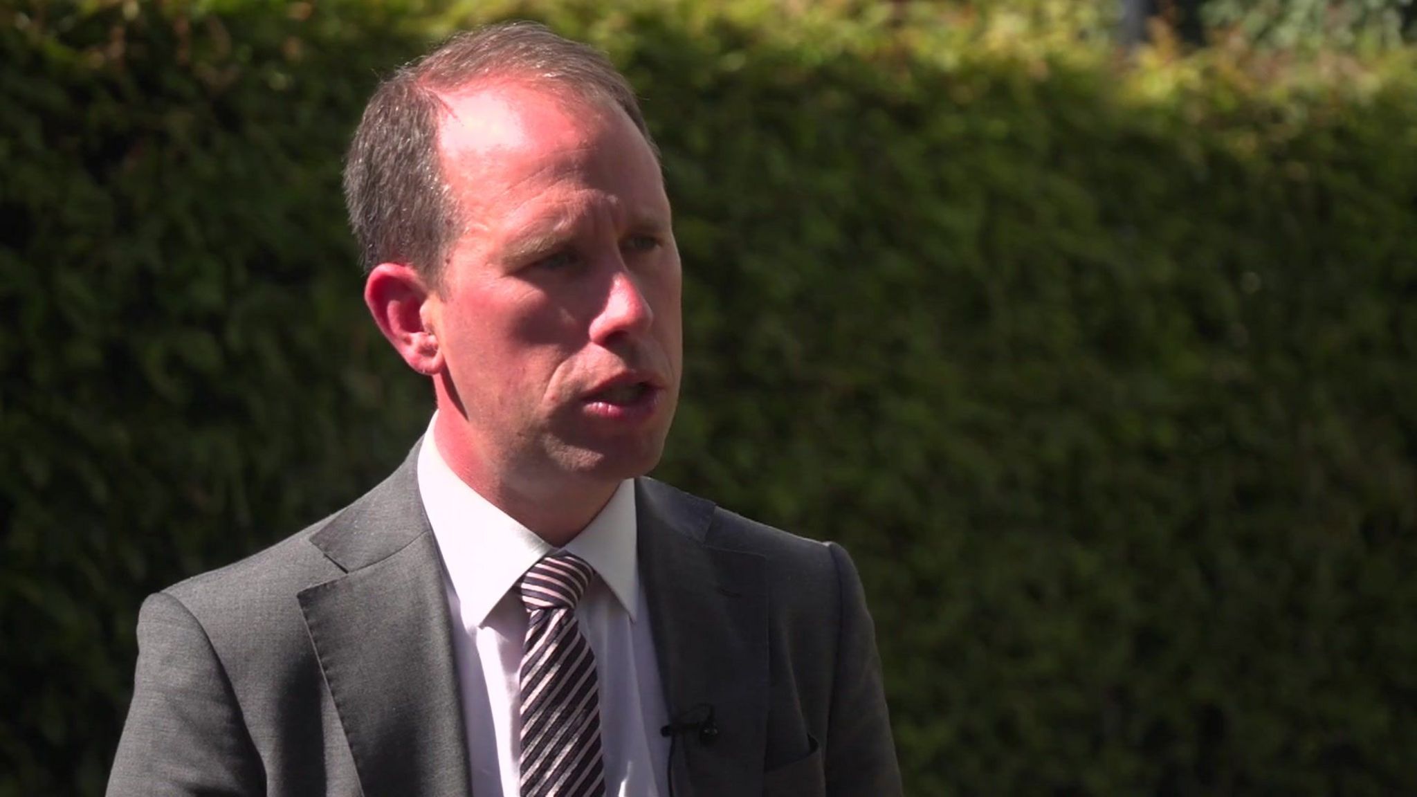 Matthew Barber. He is wearing a suit and visible from the sternum upwards. In the background is a green bush