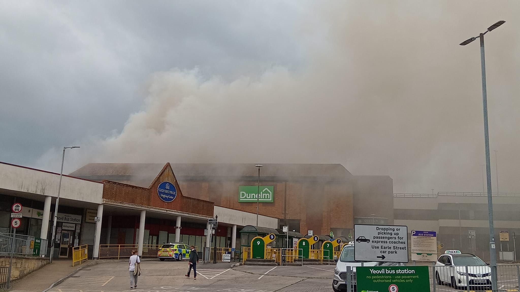 A Dunelm store in the background with a shopping centre entrance and bus station in the front and a huge plume of smoke over the scene