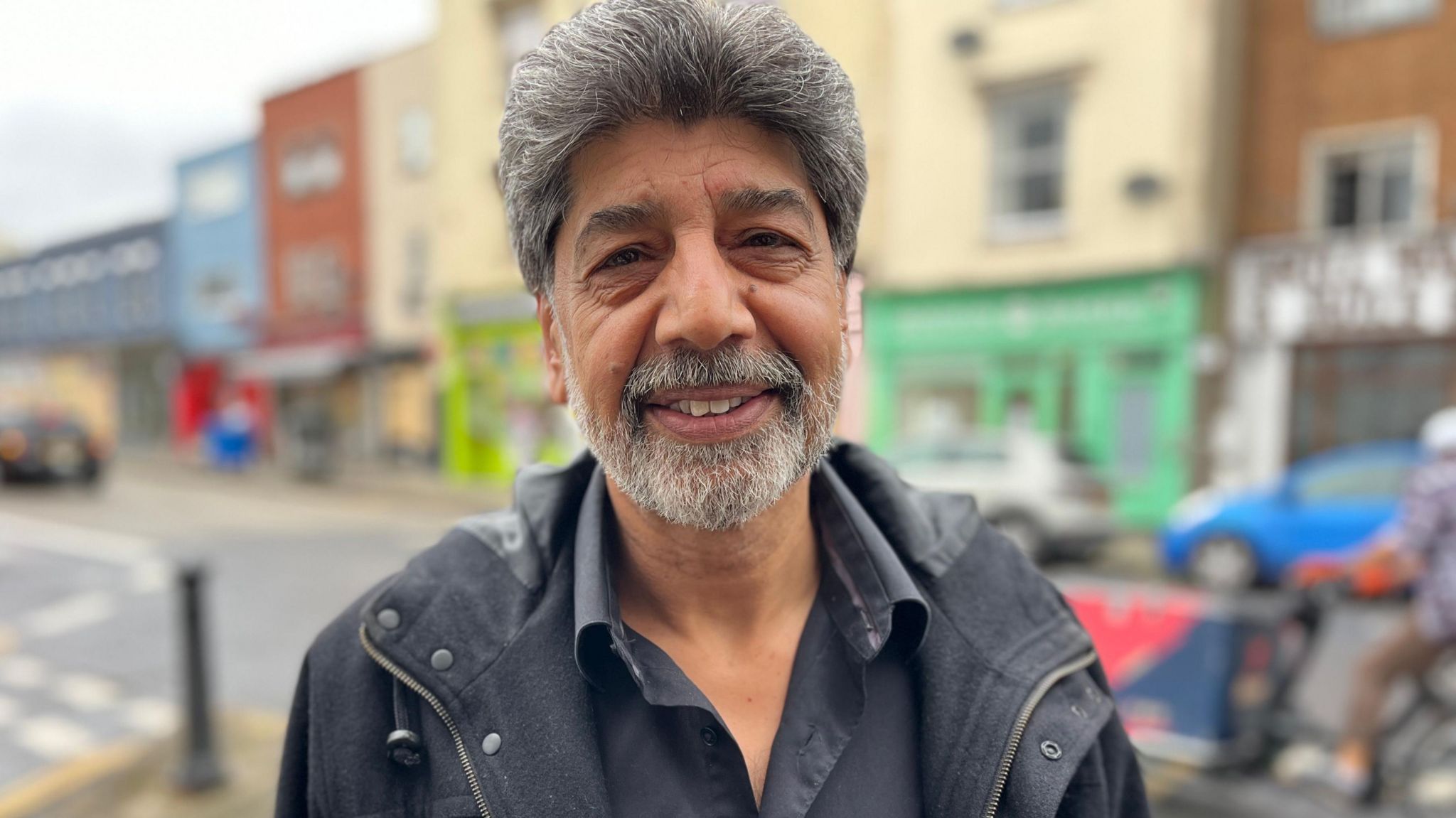 A man with a bid wearing a black jacket standing in front of a road