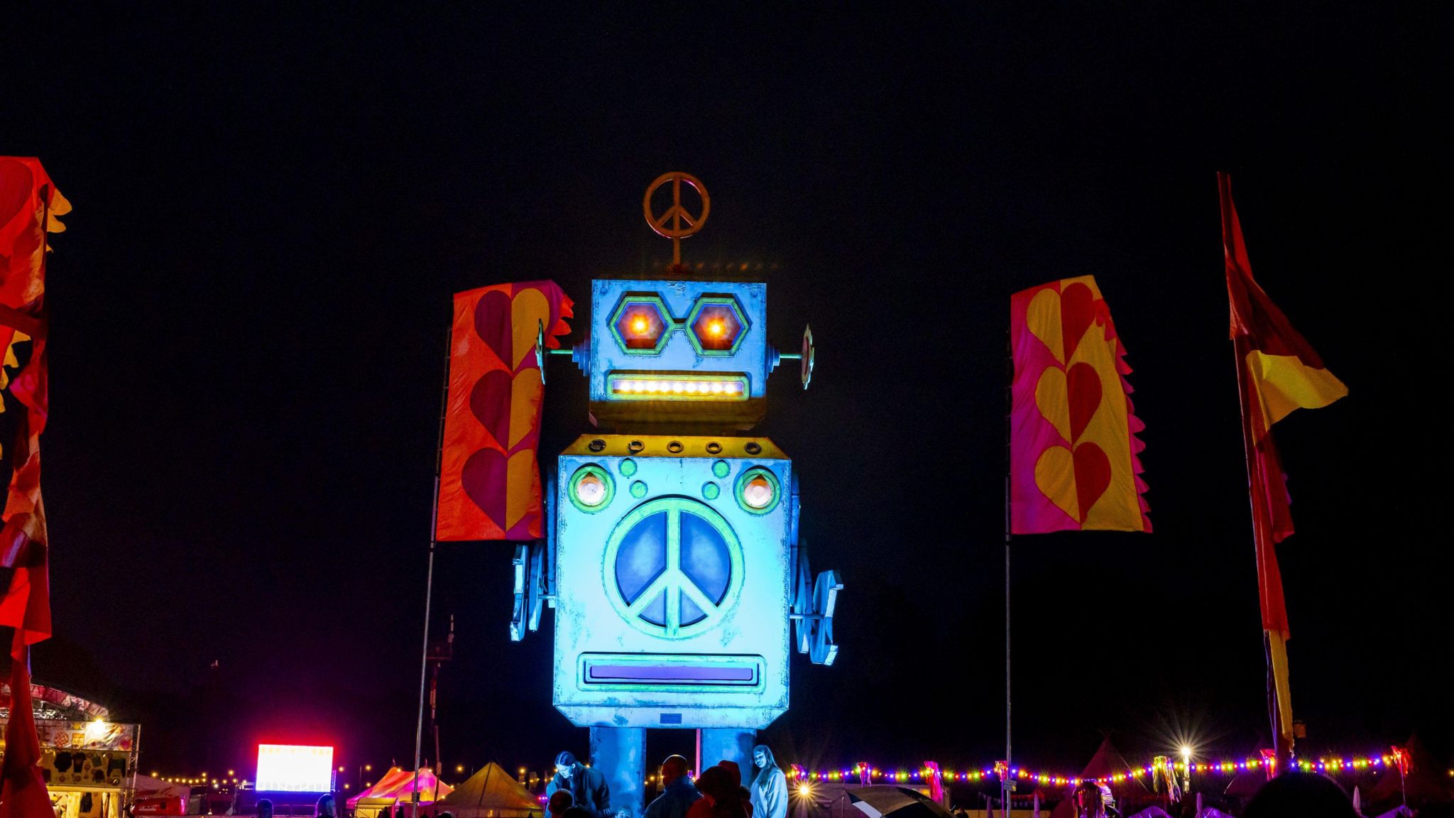 A huge blue robot stands, lit up with the night sky behind it. It has a peace sign on its head like an aerial, and a peace sign on its body. It has orange eyes. Around it, are large pink and yellow flagpoles