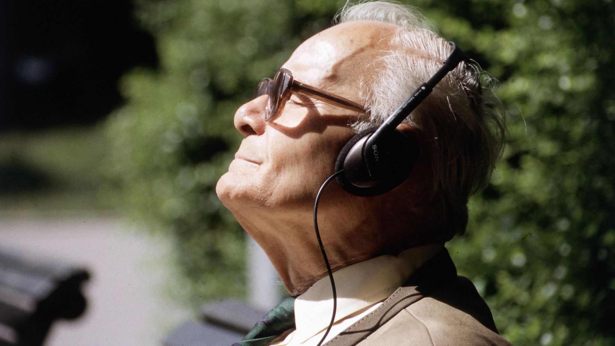 A man with grey hair and glasses smiling while listening to music with headphones on. He is outside. There are benches and greenery behind him. It is a sunny day.