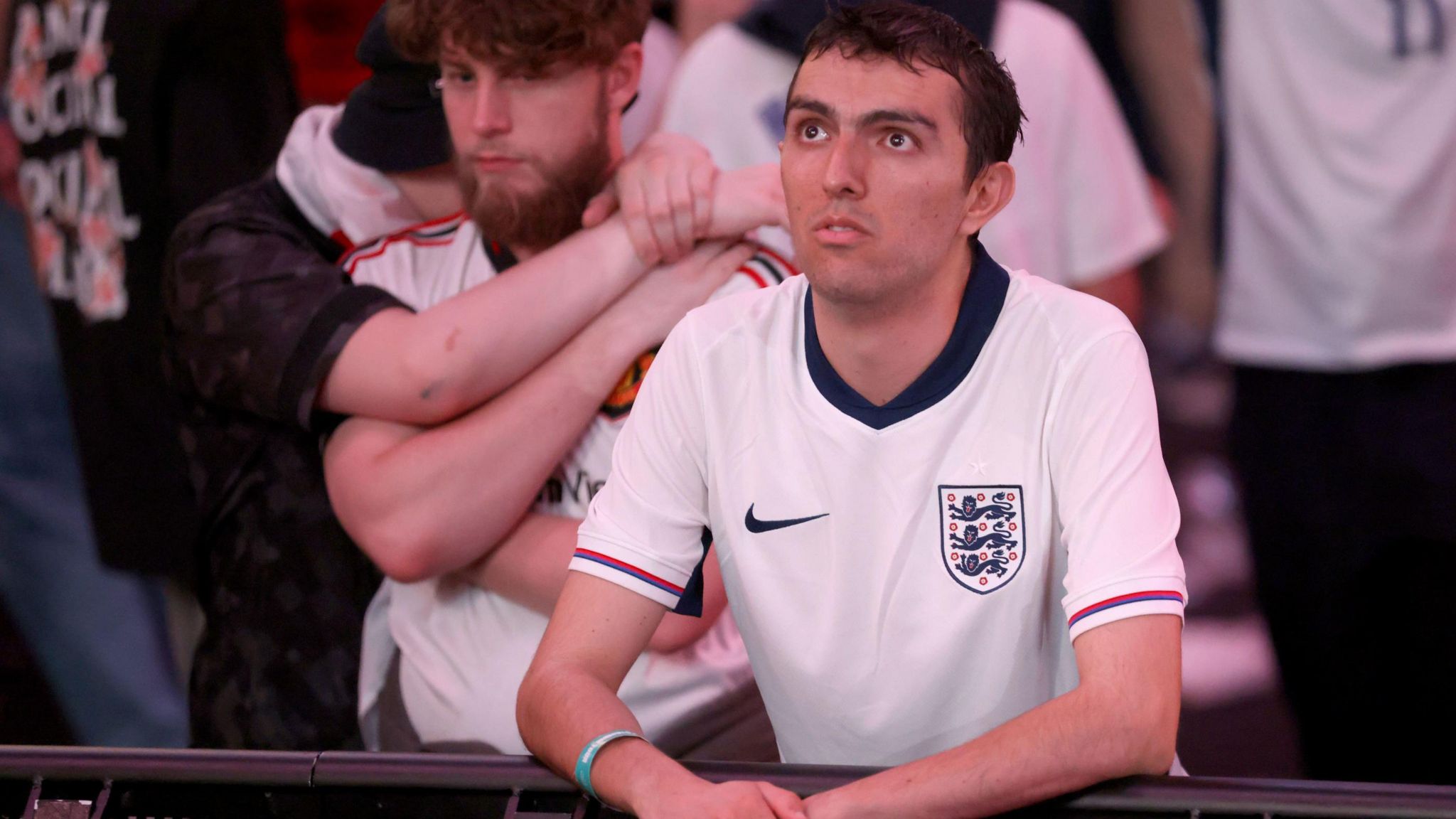 A fan wearing an England shirt leans on a barrier and stares up at a screen, while two other fans console each other