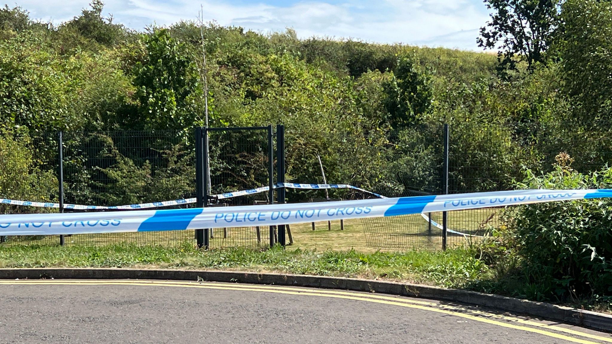 Blue and white police tape can be seen in the foreground of the picture, with a hole visible through a high chain-link fence in the distance. 