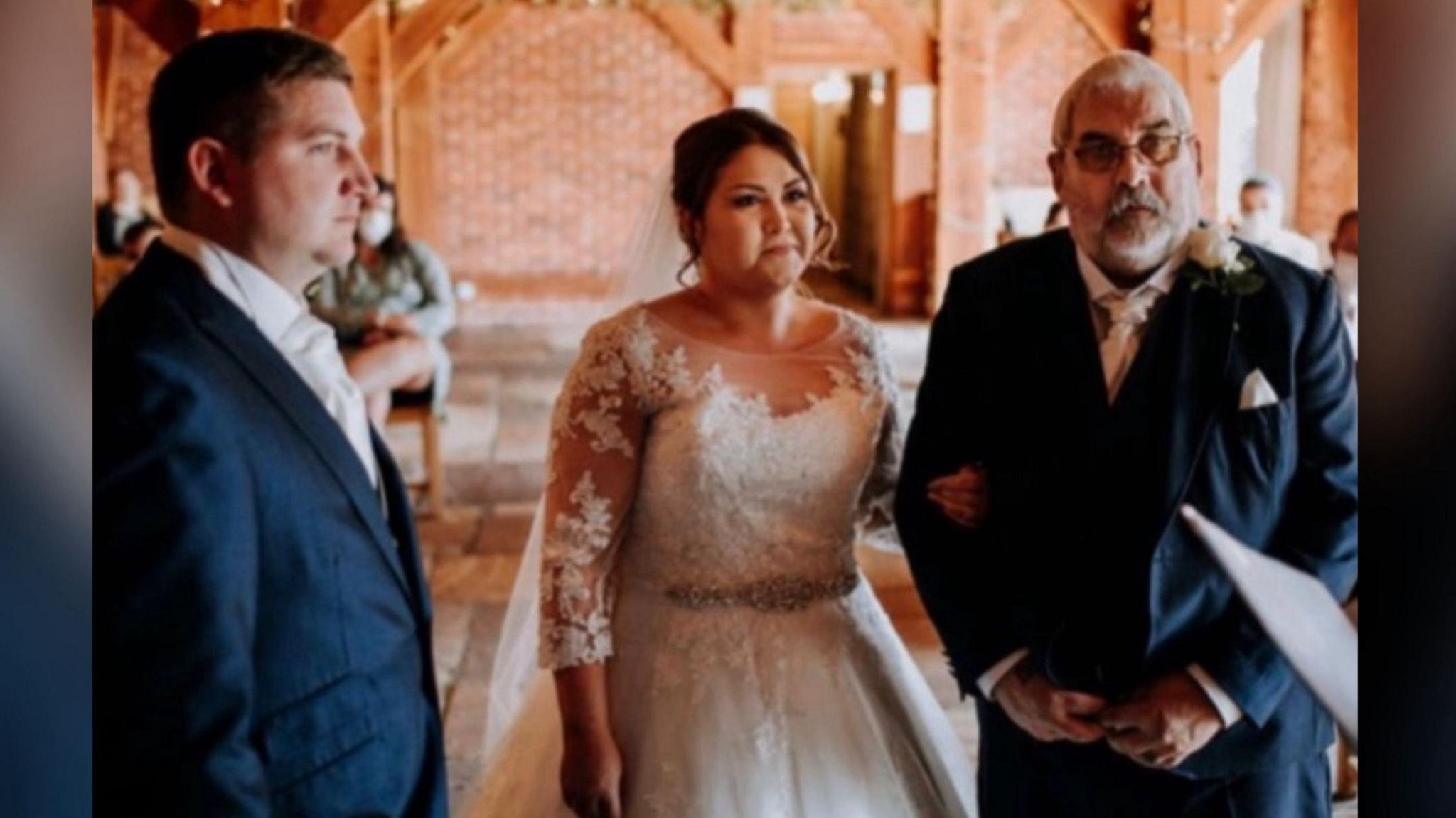 A woman in a long-sleeved white lace wedding dress stands between her husband and father in navy blue suits on her wedding day