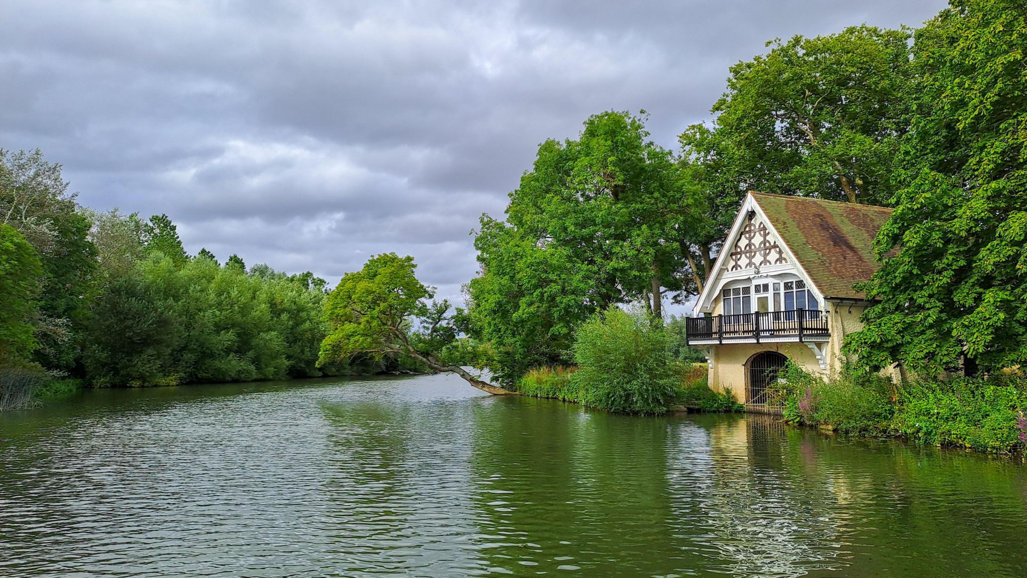 A cream and white boat house with a pitched roof pokes out amongst trees from the right of the image. It has a balcony and a gated entrance at water level. The river reflects the green of the trees either side of it and the cloudy skies above.