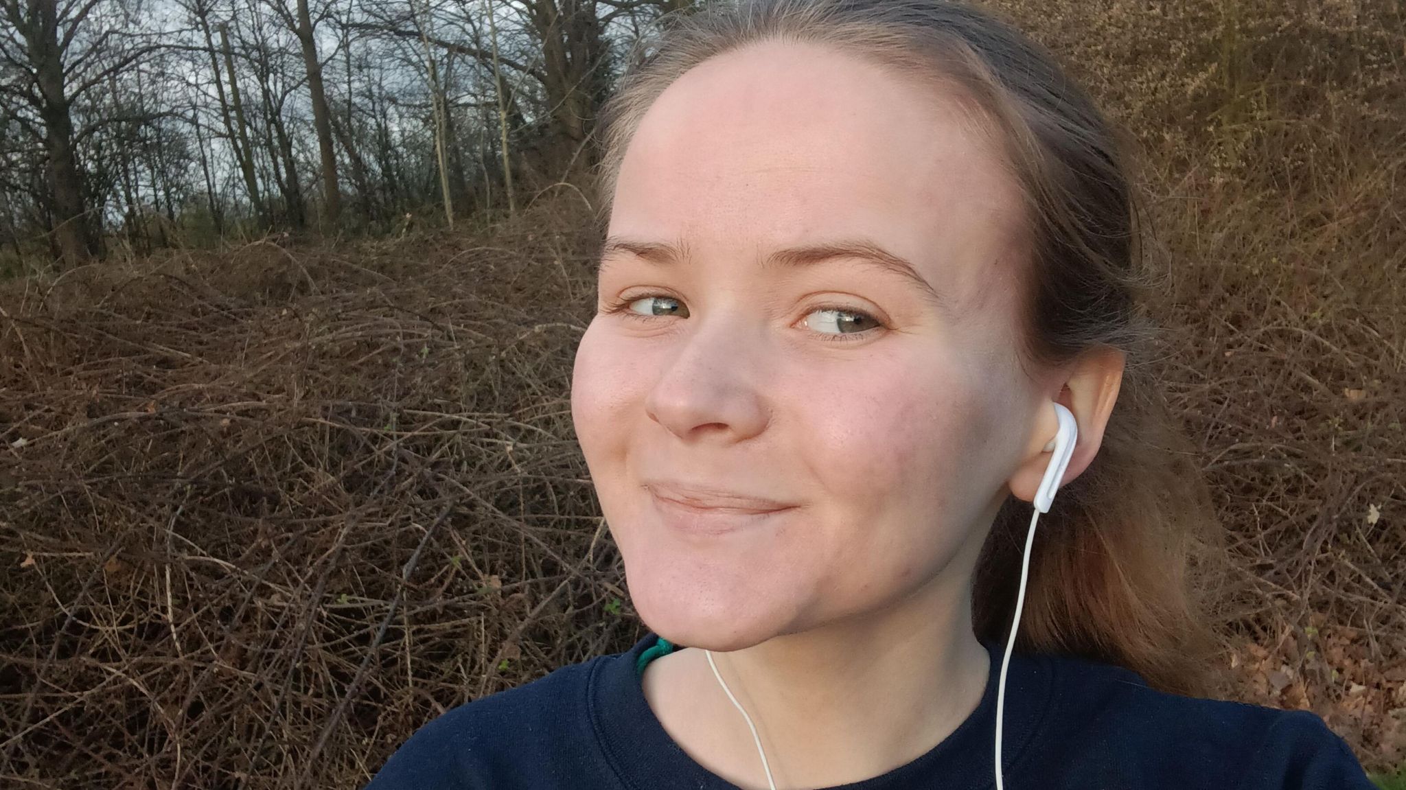 Chloe has clearer skin and her light brown hair is up in a ponytail . She is wearing a navy jumper and white in-ear headphones