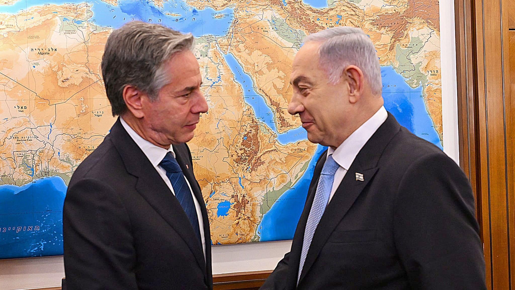 Blinken and Netanyahu shaking hands during a recent visit. The two men are standing front of a world map and look serious. 