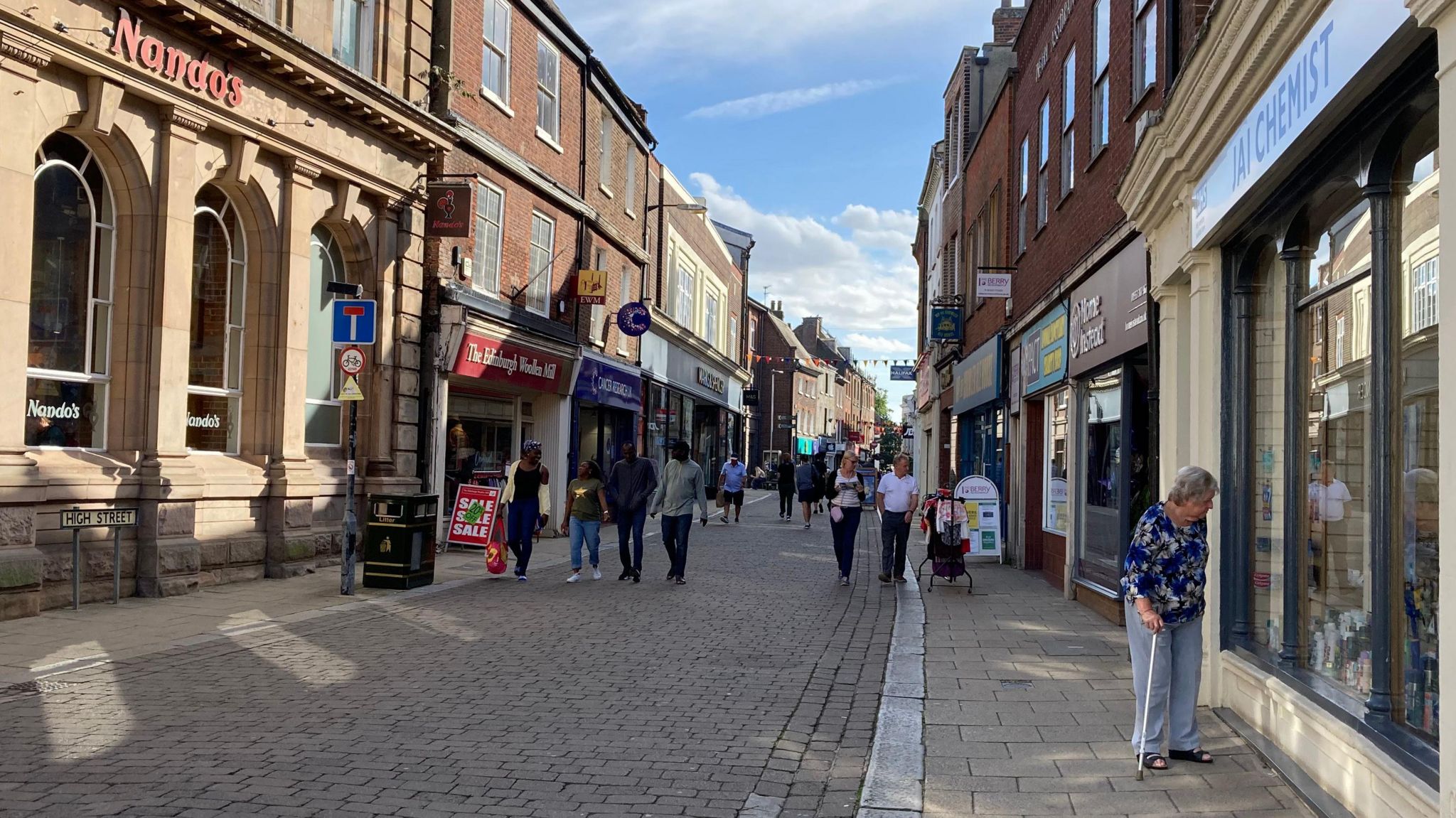 A street in King's Lynn: A cobbled pavement lined with two flagstone walking paths on either side next to shops. A woman in a floral top, with a walking stick is looking in a window in the foreground and groups of people are walking with bags in the background