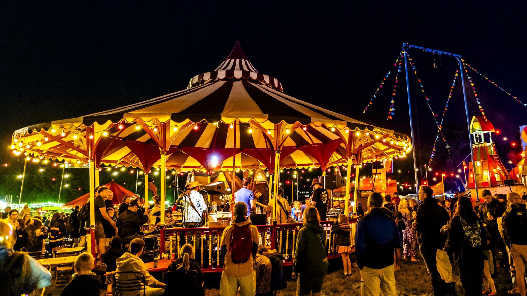 A carousel-style tent with red and white stripes is covered with yellow lights. Dozens of people surround it, watching a musical act inside. In the background are colourful bunting strings, attached to a tall blue structure