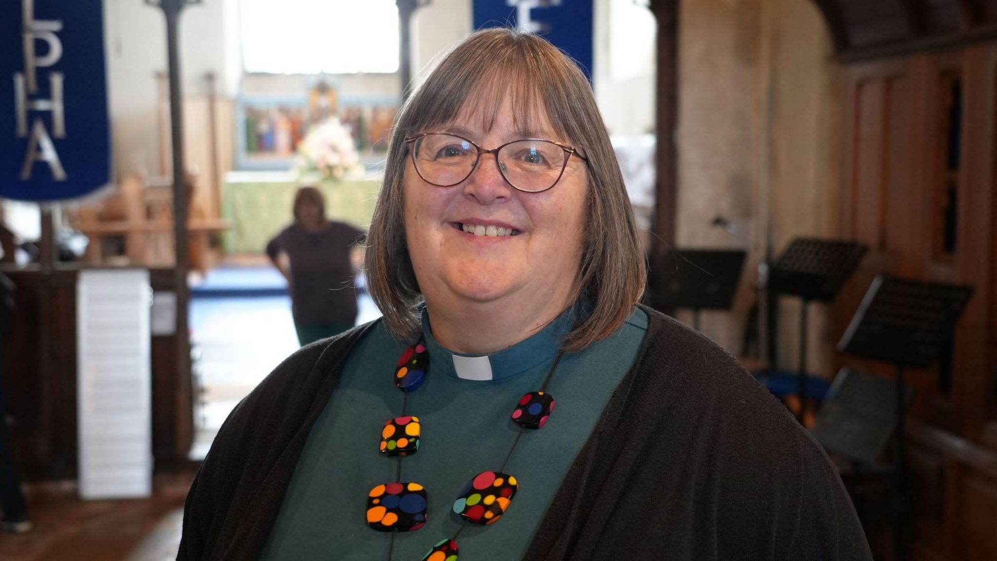 Veronica Wilson wears a green cassock and dog collar, and around her neck is a brightly coloured necklace. She  stands in a church. She is also wearing spectacles.