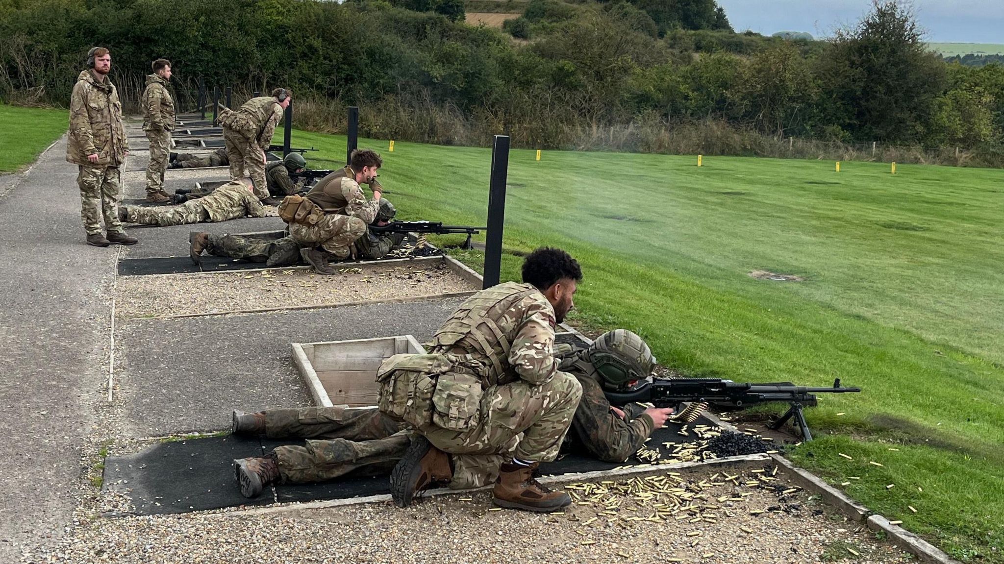 A row of soldiers lying in the prone position shooting a machine gun with more soldiers kneeling over them.