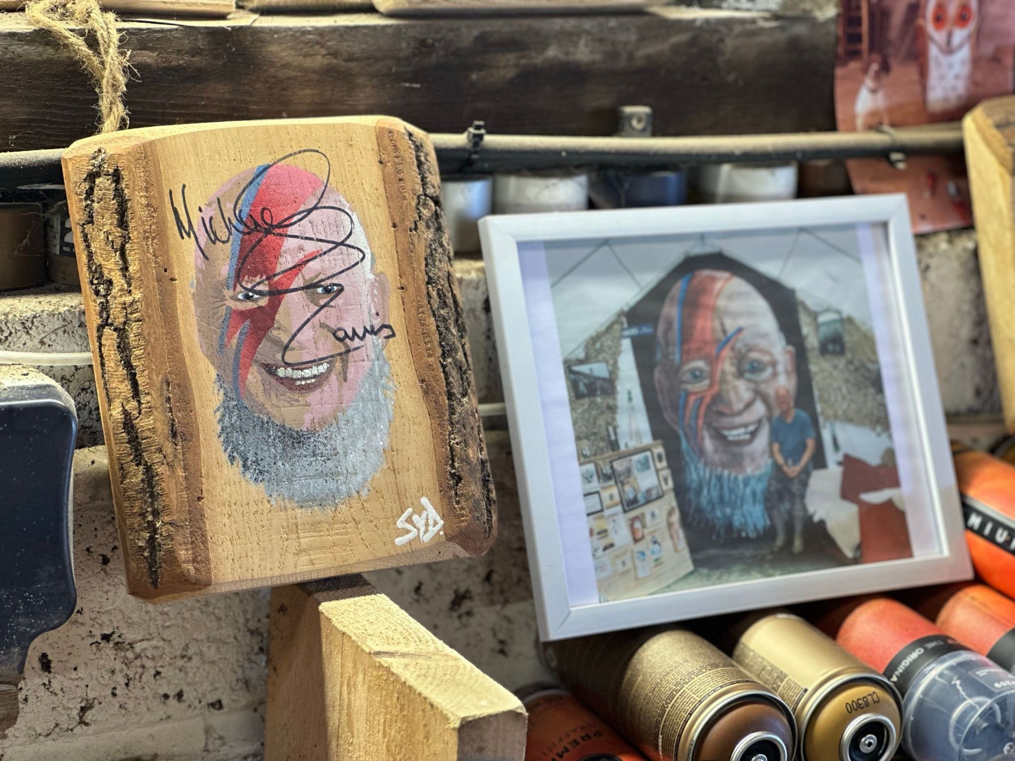 A wooden block with Michael Eavis' face, signed by him and a framed picture of the artist standing next to a large mural of the same image