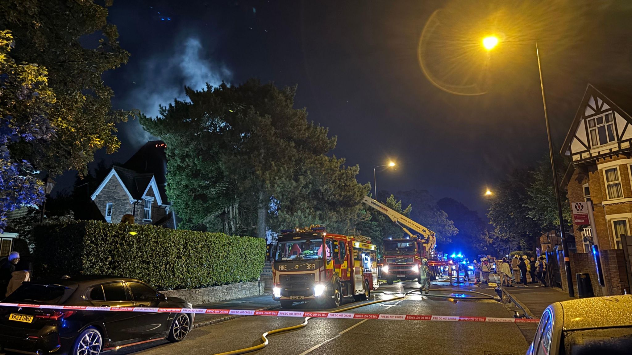 Several fire engines can be seen parked in a residential street at night, with smoke rising up in the distance above a tree